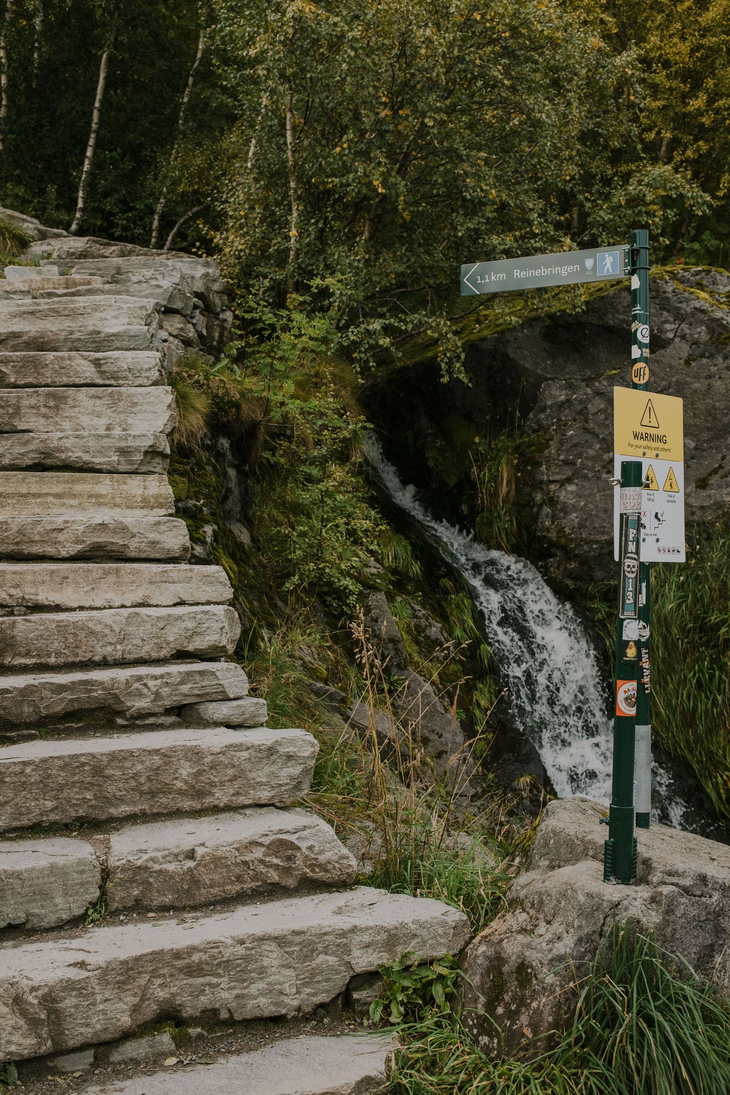Stone steps lead up alongside a waterfall with hiking trail markers in a lush forest setting.