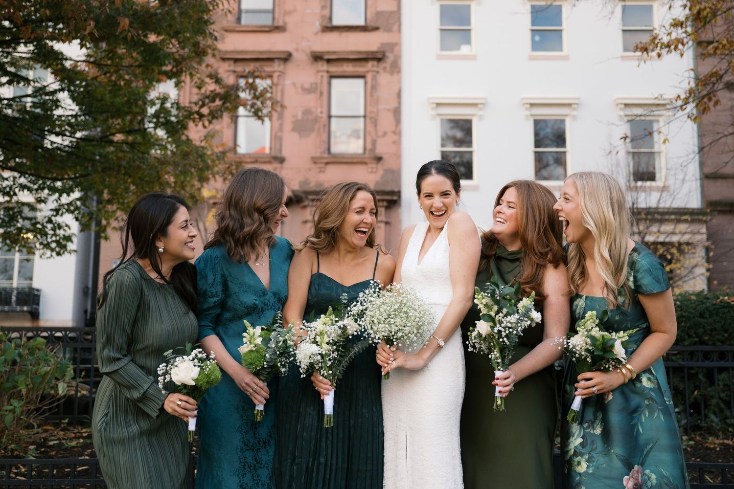 Wedding party in emerald green dresses standing together with bouquets outside brownstone buildings.