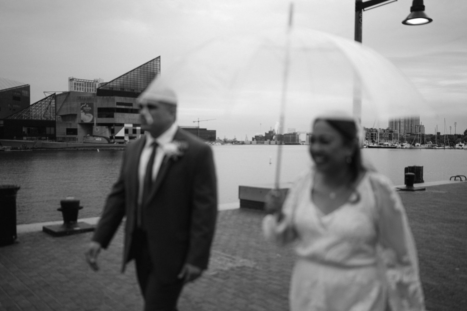 A black and white photo shows a couple walking along a harbor boardwalk with a white parasol.