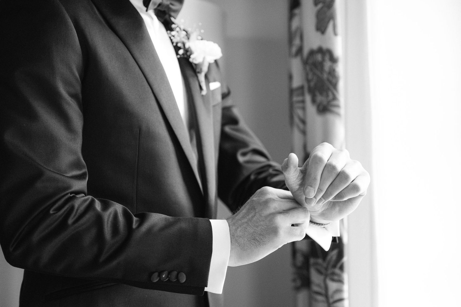 Black and white photo of hands adjusting a crisp white boutonniere against a formal black suit jacket.