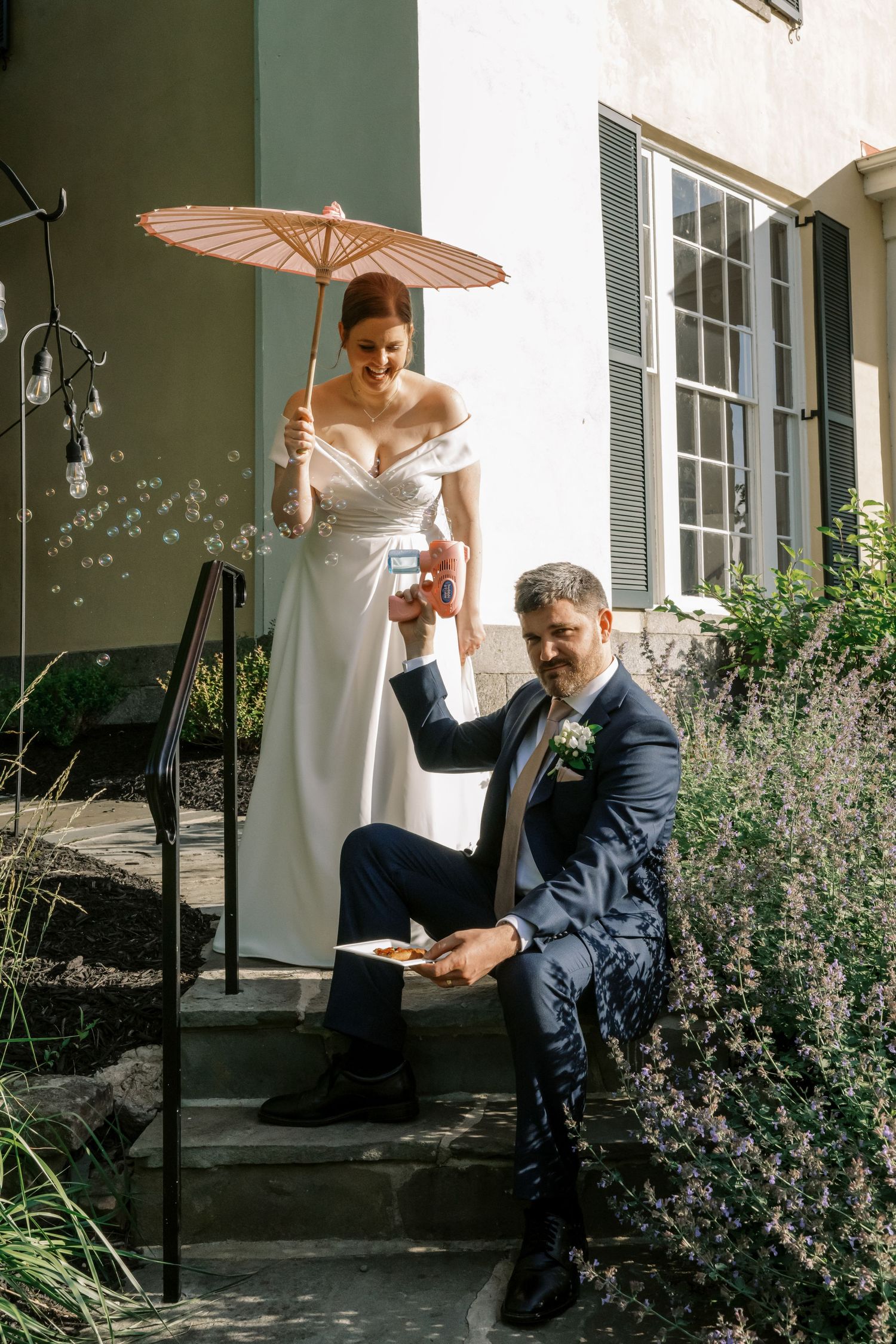 A couple poses on stone steps with a pink parasol and bubbles floating in the sunlight.