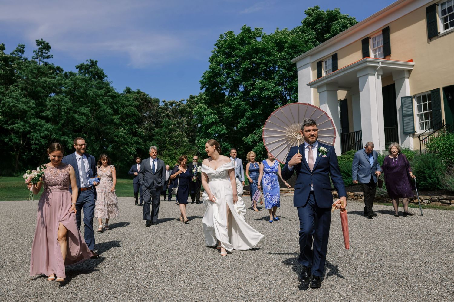 Wedding party walking outside stately venue on gravel path surrounded by greenery.
