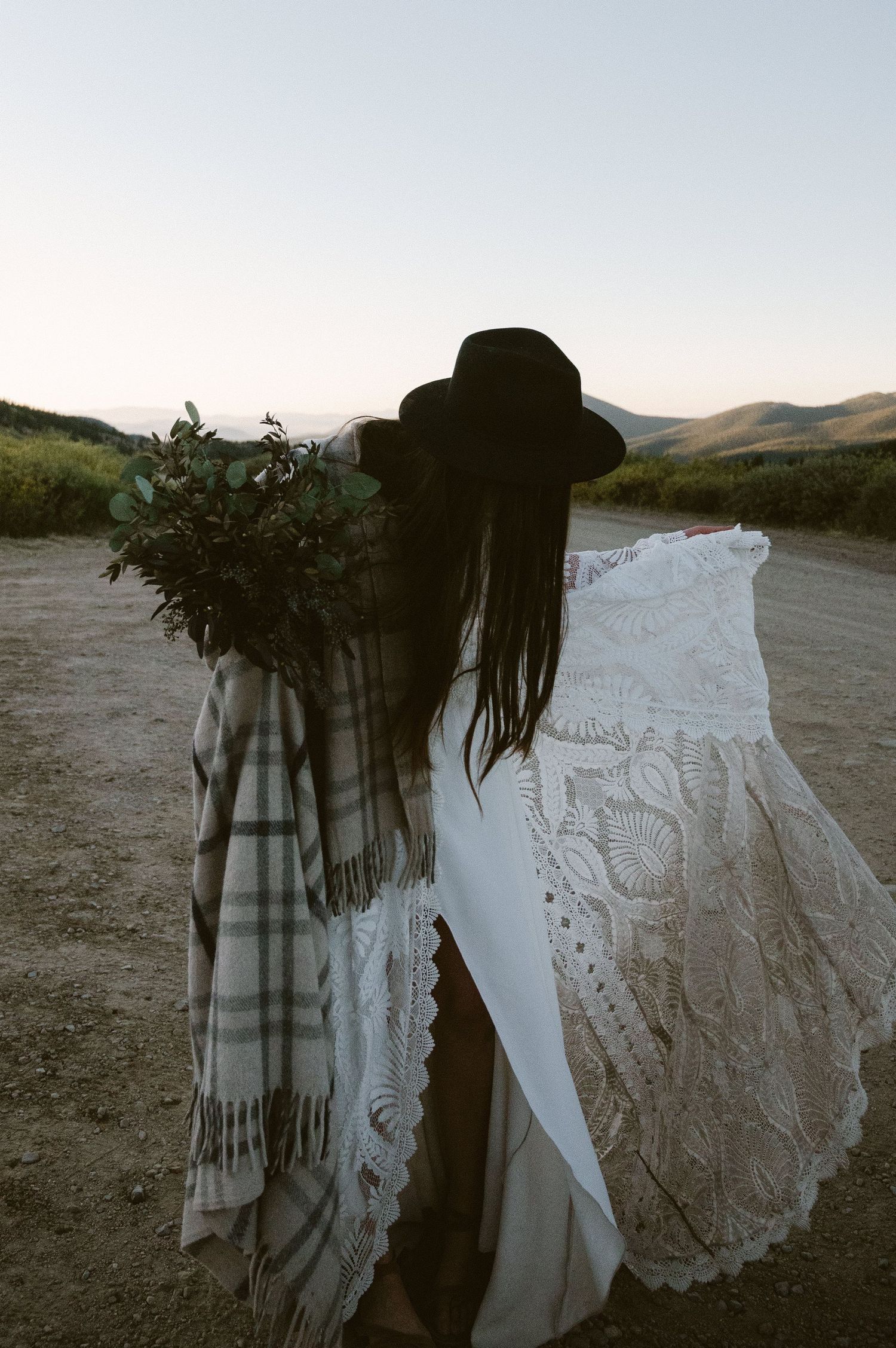 A figure in a white lace dress and hat holds a bouquet against a mountain backdrop.
