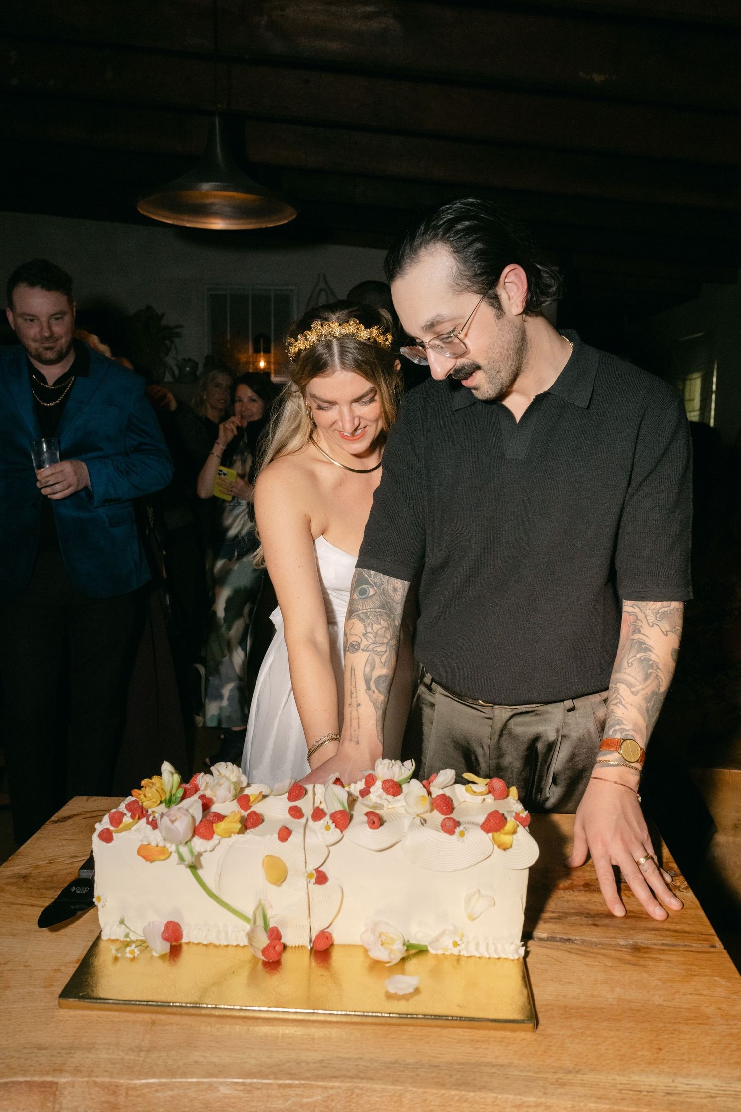 Two people in a dimly lit room work together to cut and serve slices from a large rectangular dessert on a wooden table.