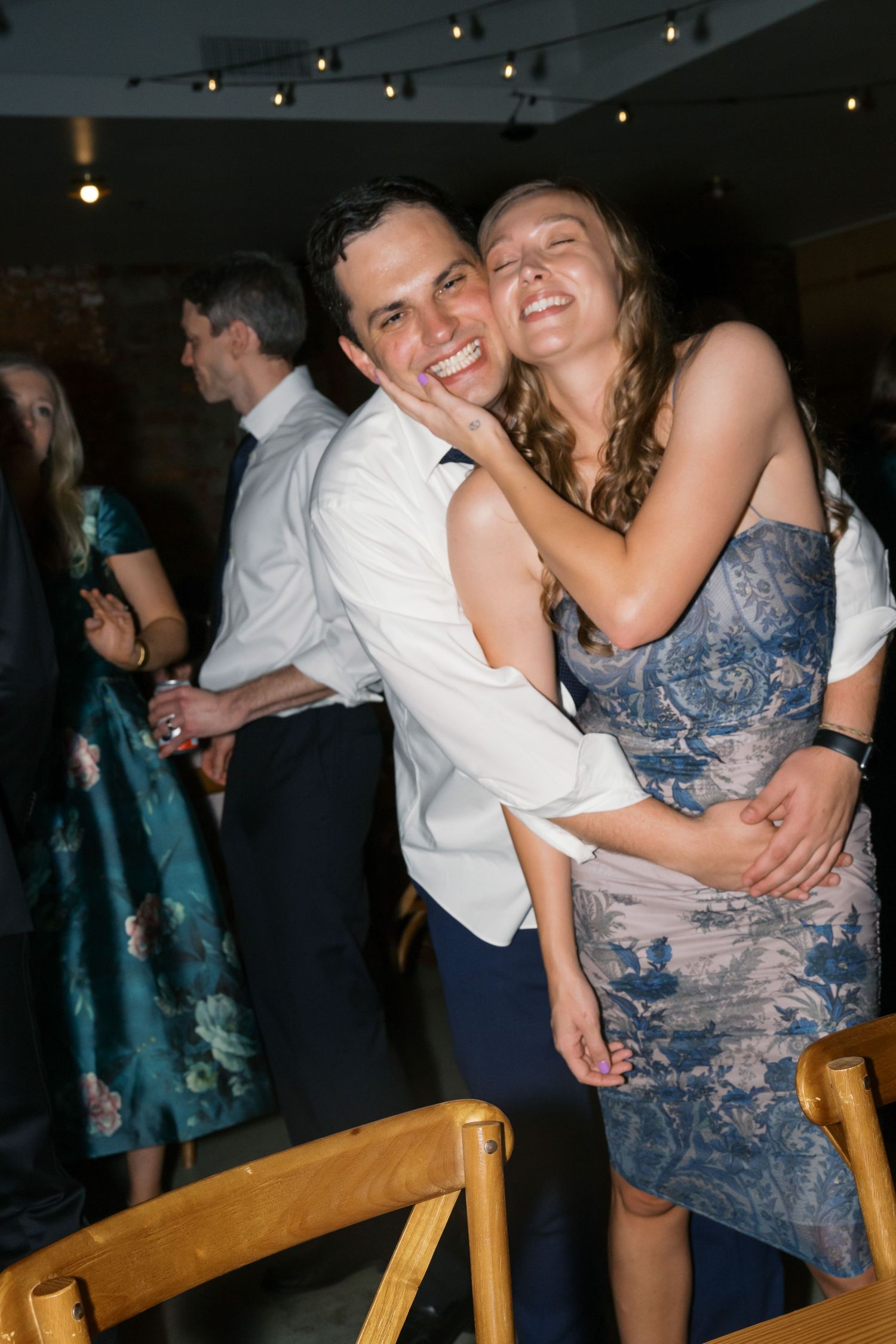 A joyful couple shares an intimate dance moment at a wedding reception, surrounded by string lights and other guests.
