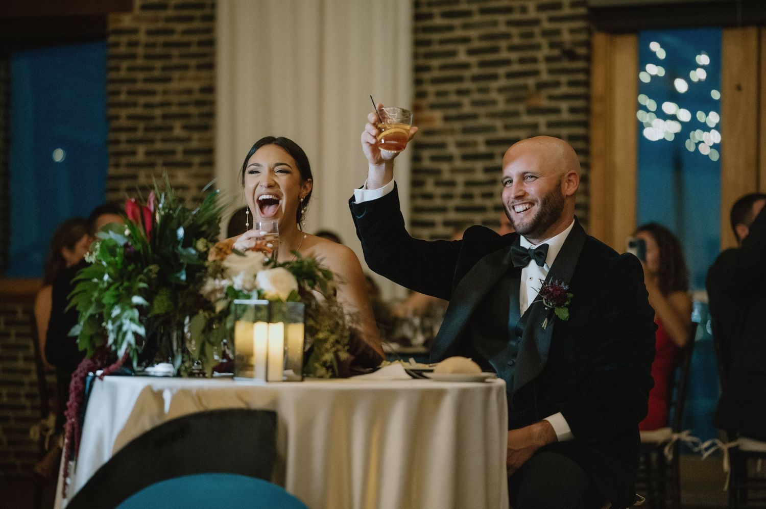 Newlyweds raise their glasses in celebration at their reception table decorated with candles and flowers.