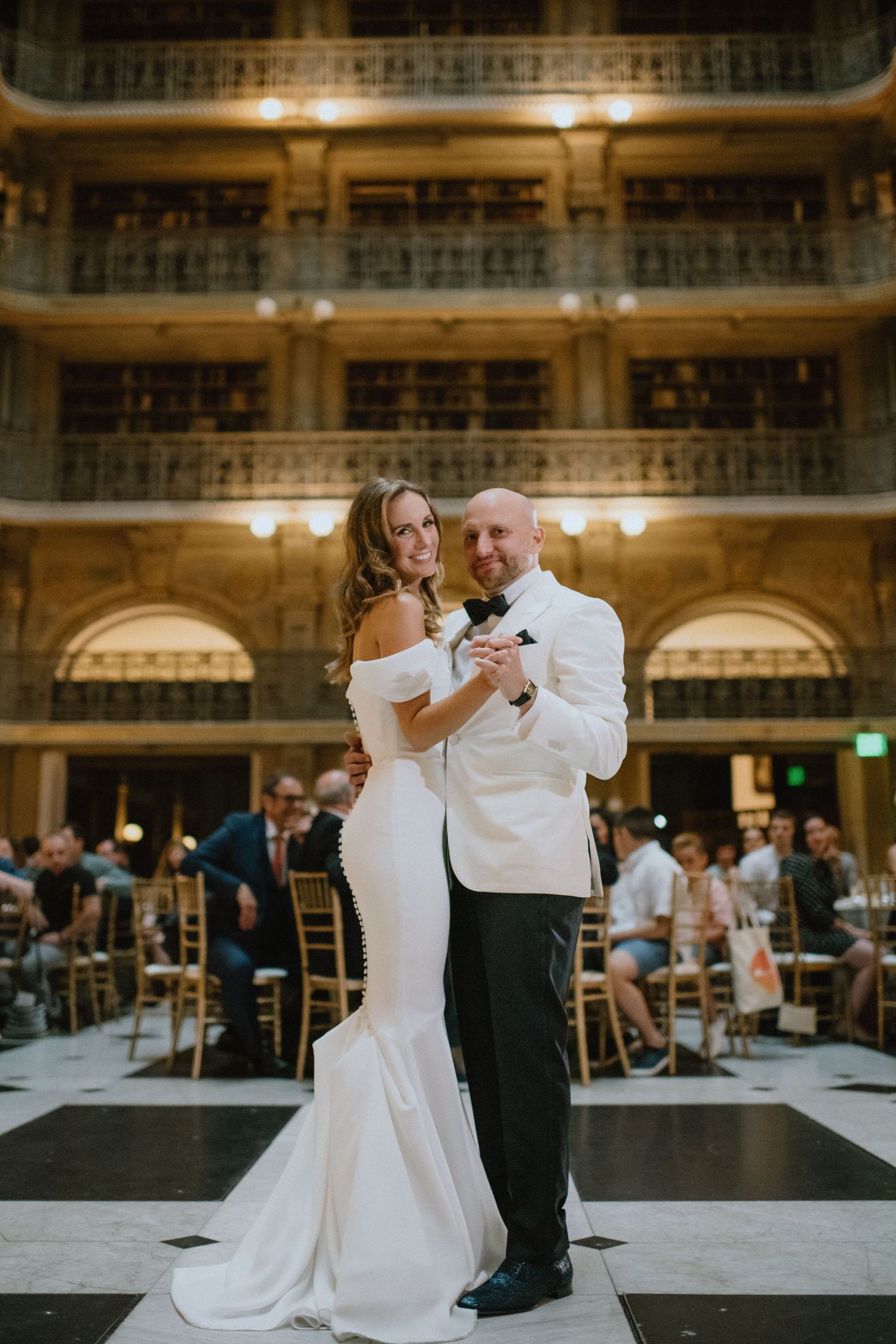 Wedding couple dancing in an ornate library with checkerboard floor and tiered balconies.