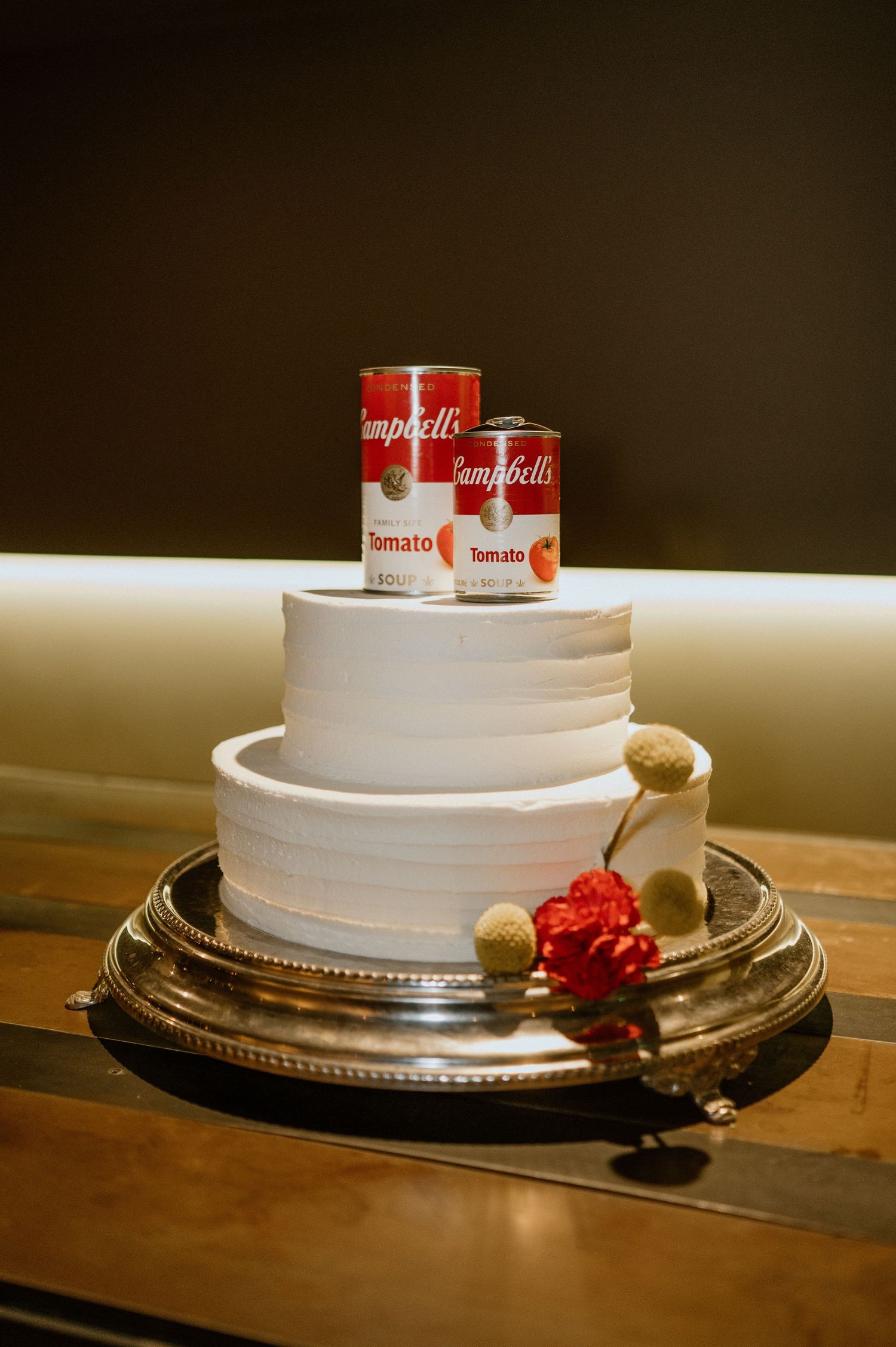 White wedding cake topped with Campbell's soup cans and red carnations on a silver platter.