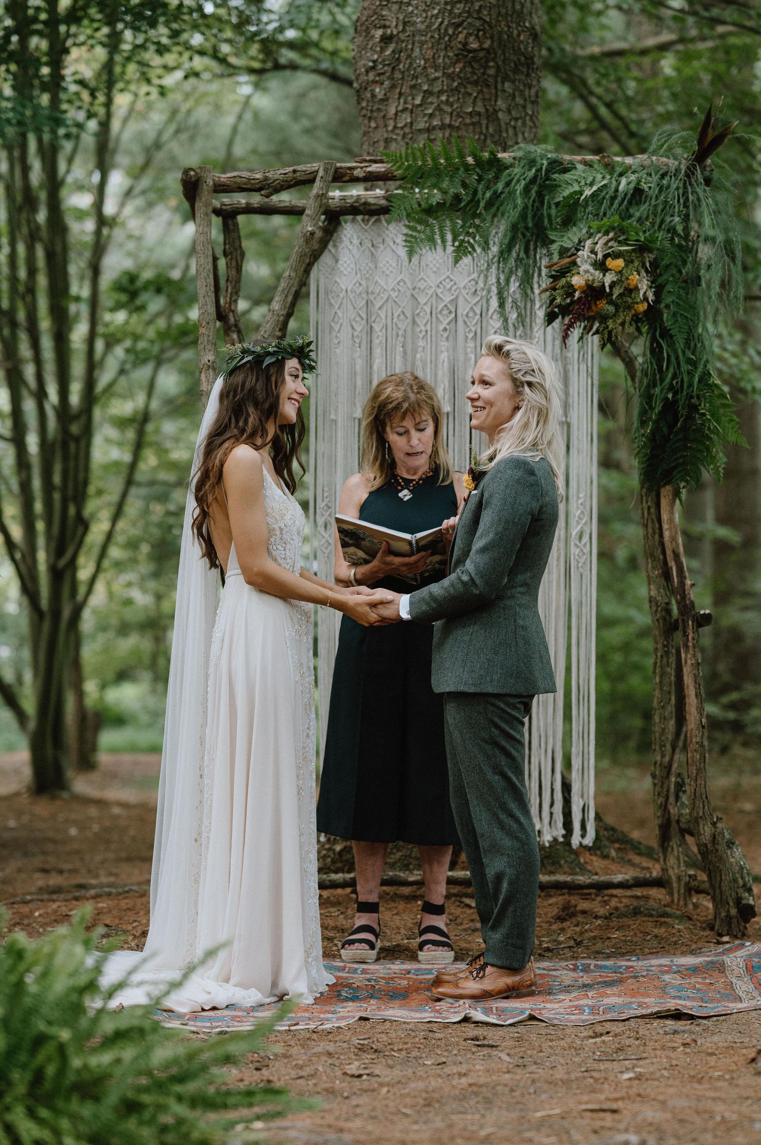 Wedding ceremony takes place under a rustic wooden arch with macrame backdrop in a forest setting.