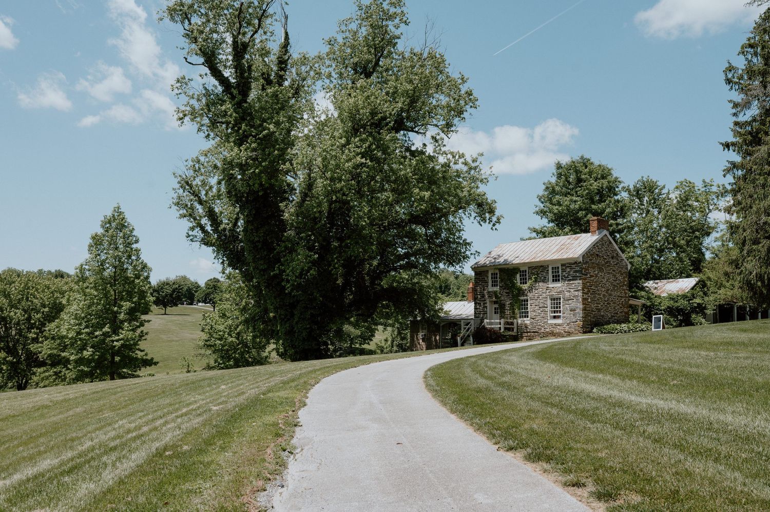 Long winding driveway leading to a rustic stone house surrounded by manicured lawn and trees.