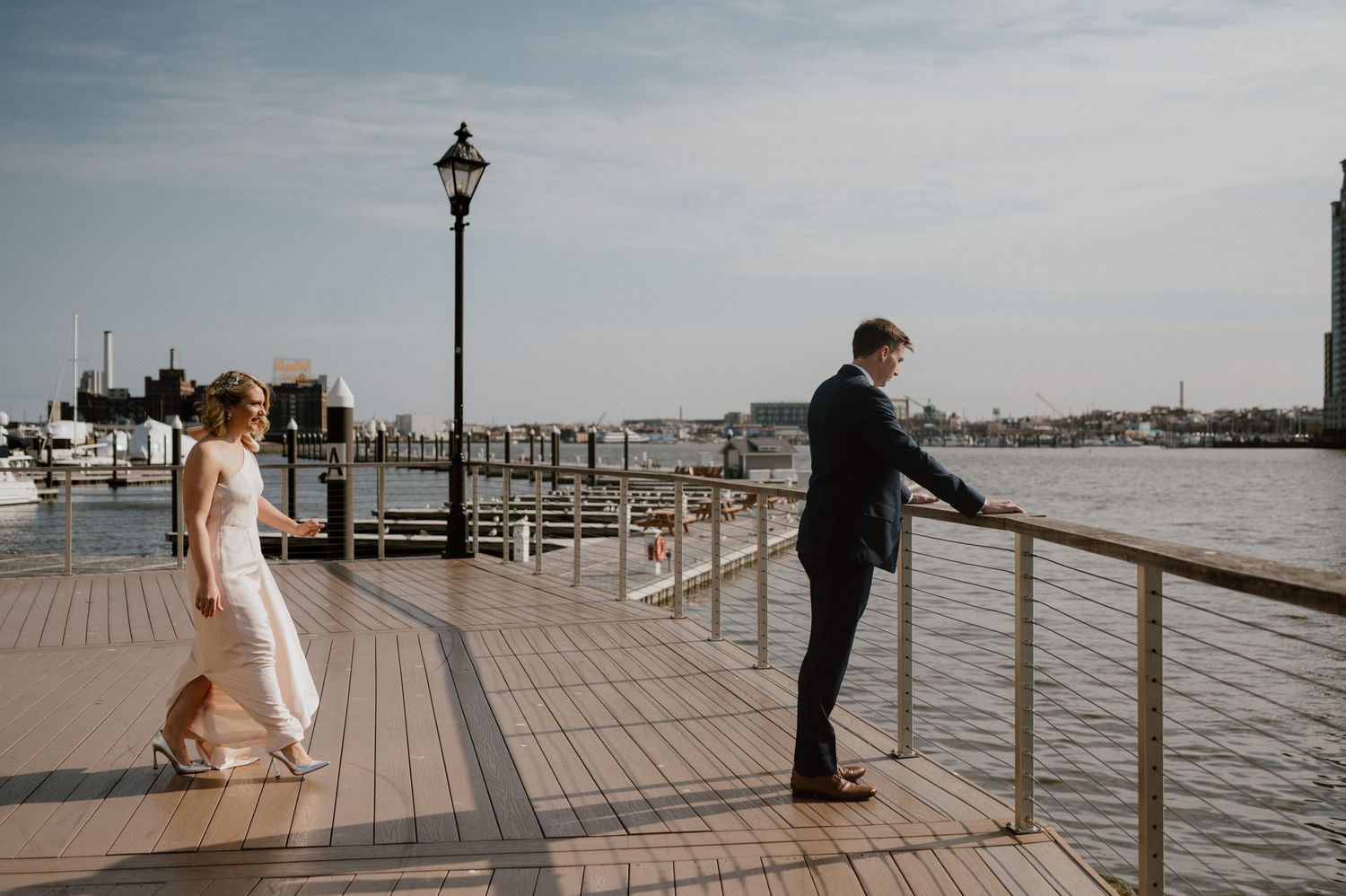 Couple stands separately on waterfront boardwalk during wedding photoshoot.