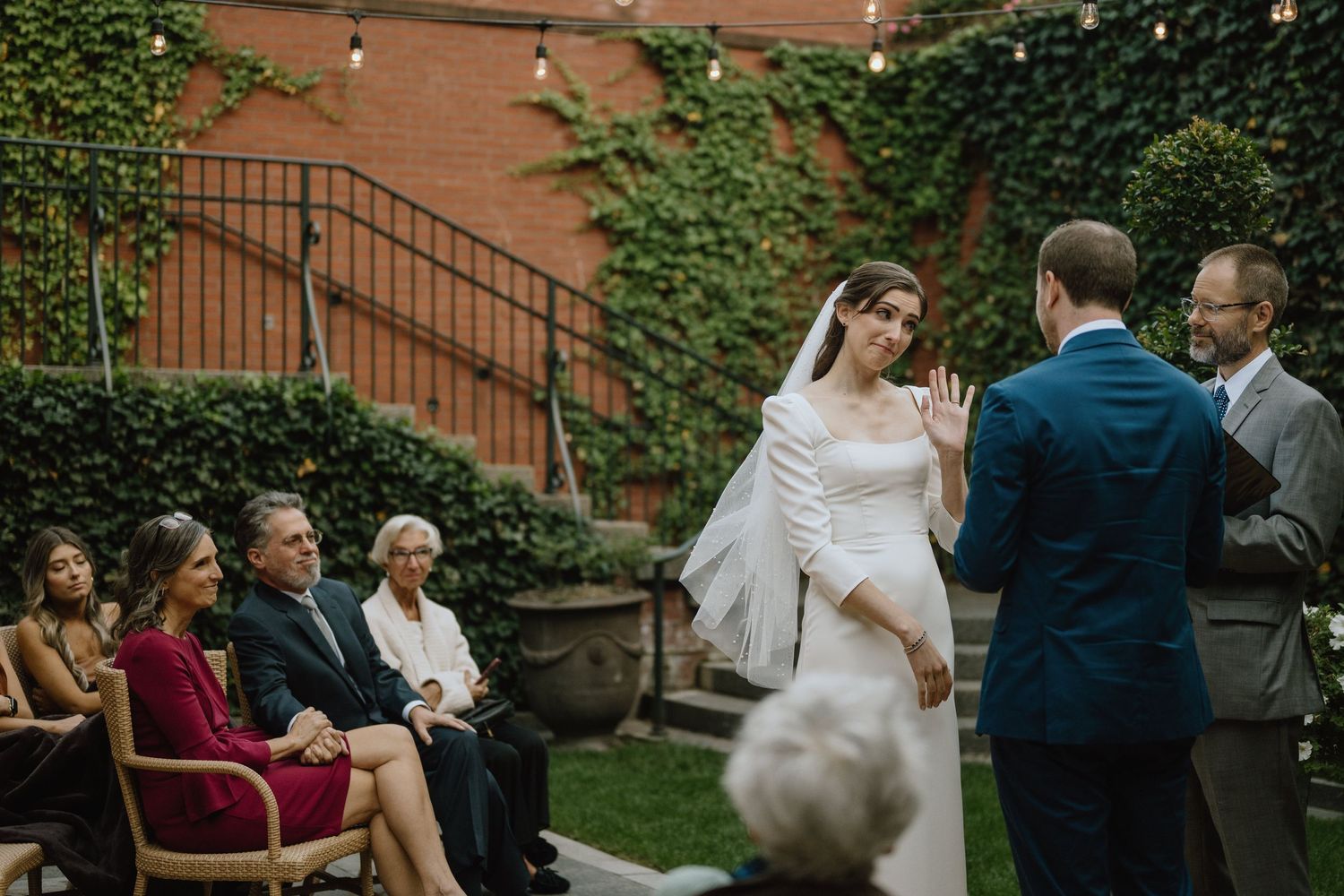 An outdoor wedding ceremony with string lights and ivy-covered walls creates a romantic atmosphere.