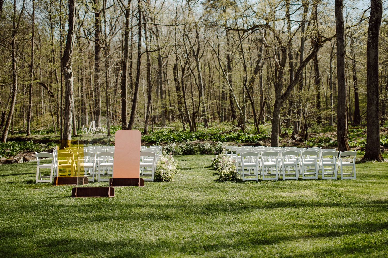 Outdoor wedding ceremony setup with white chairs and pink accents in a wooded setting.
