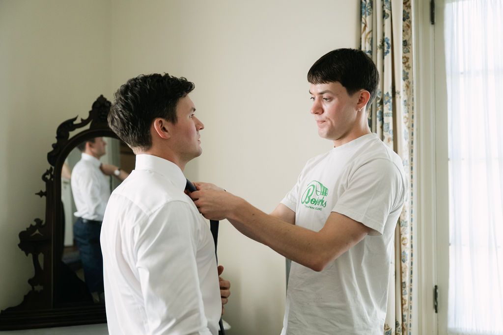 Two people in white shirts help with formal attire preparations in an elegant room.