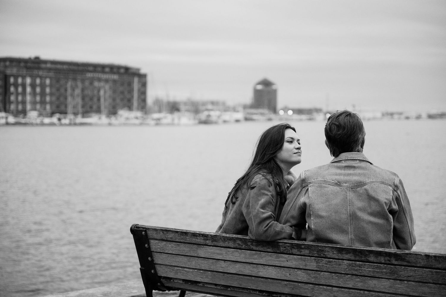 Black and white photo of a couple sitting on a bench by the waterfront, sharing a quiet moment together.