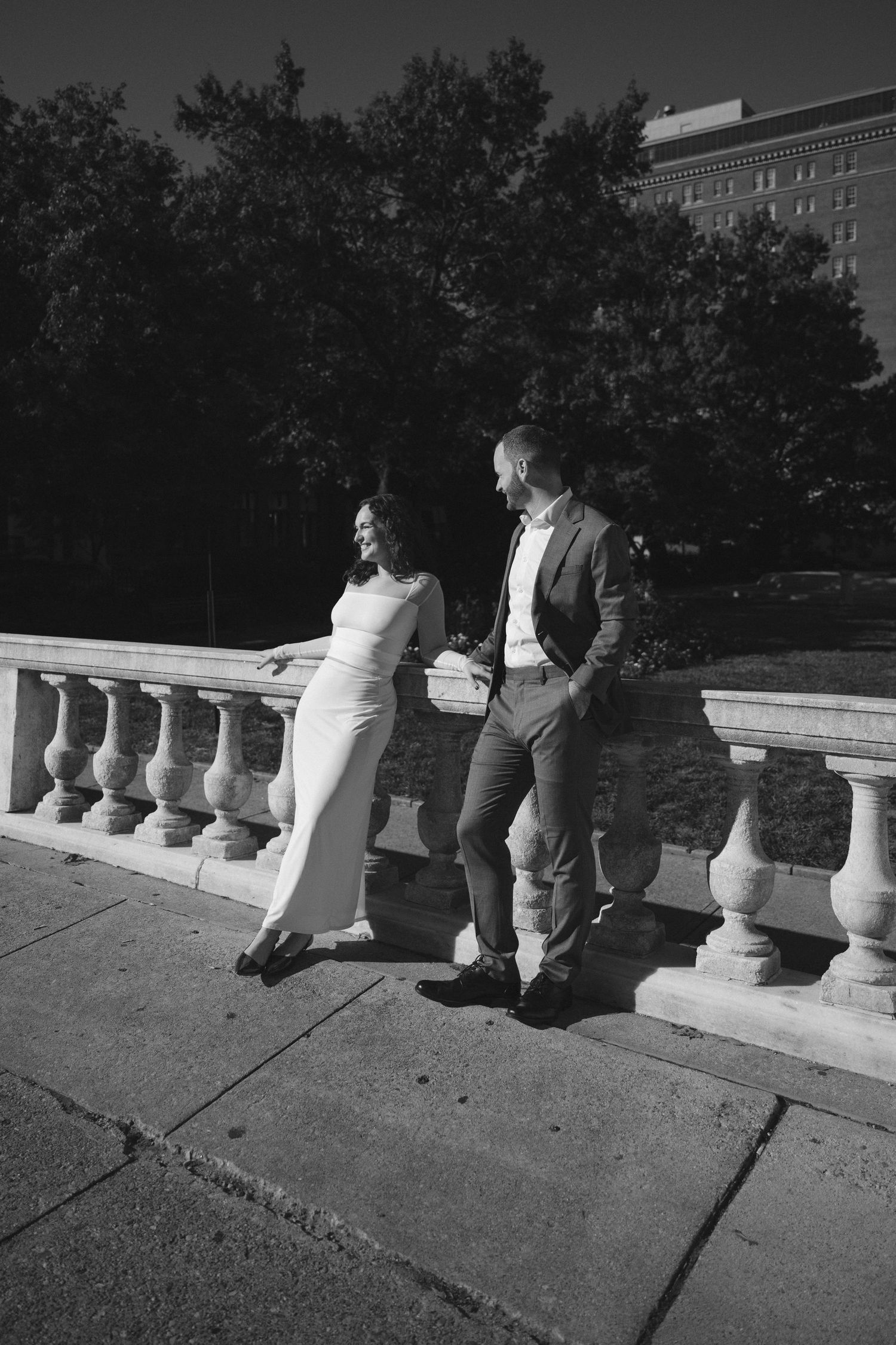 Black and white photo of a couple in formal attire standing by a stone balustrade in an urban park setting.