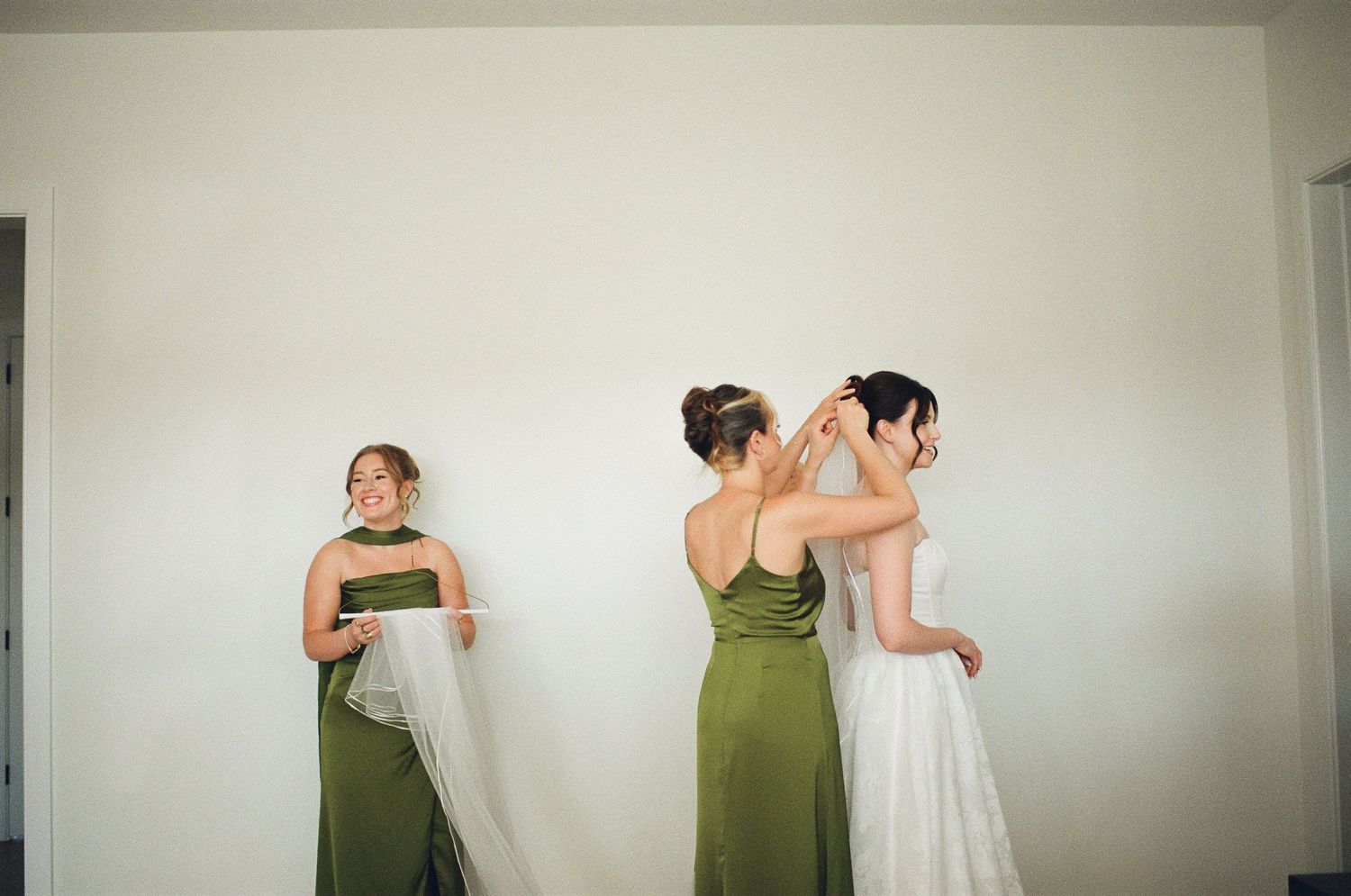 Bridesmaid in green dress helps bride with veil while another bridesmaid watches in a bright white room.