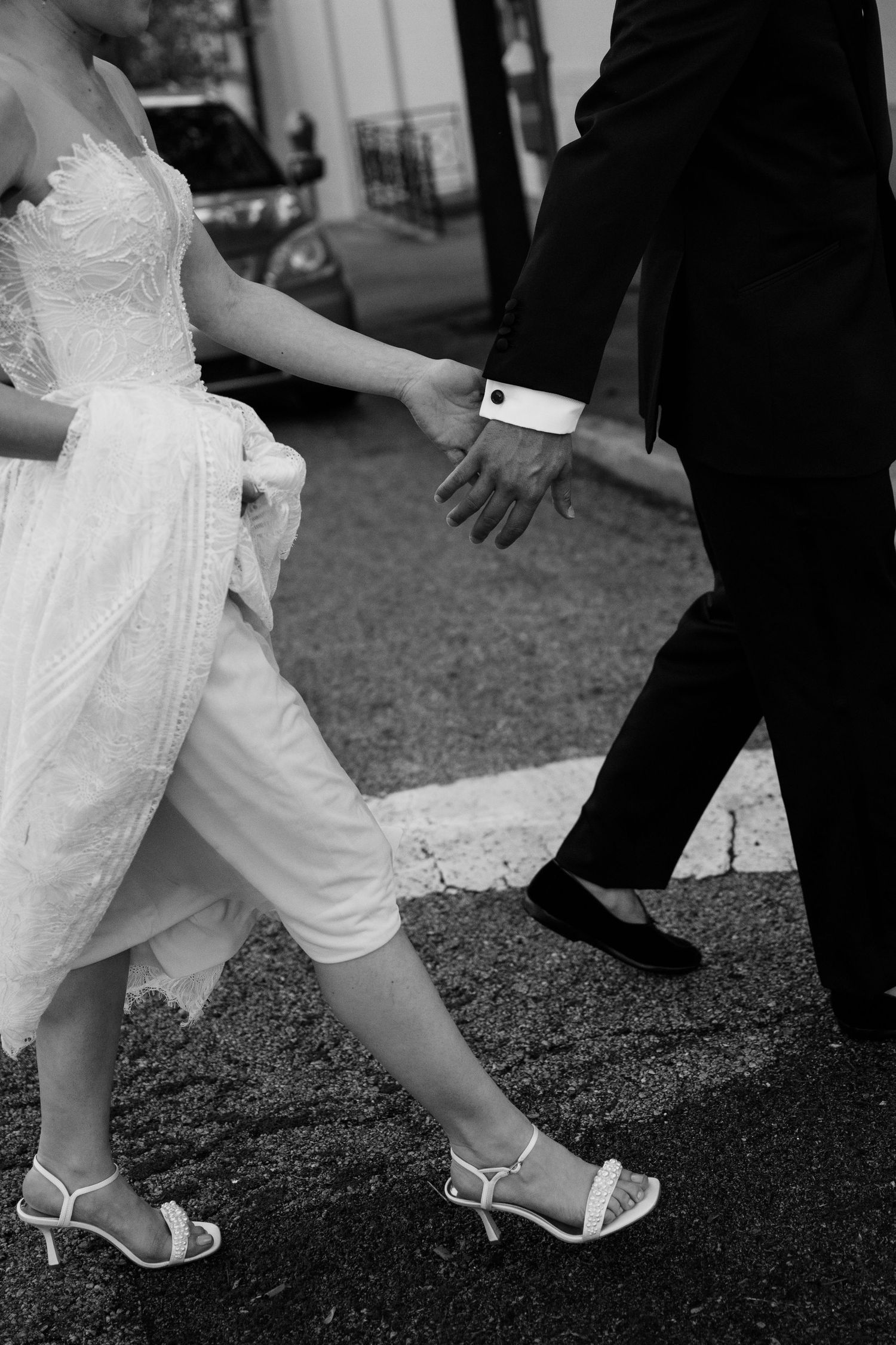 Black and white photo of bride and groom holding hands, walking in wedding attire, bride wearing white heels.