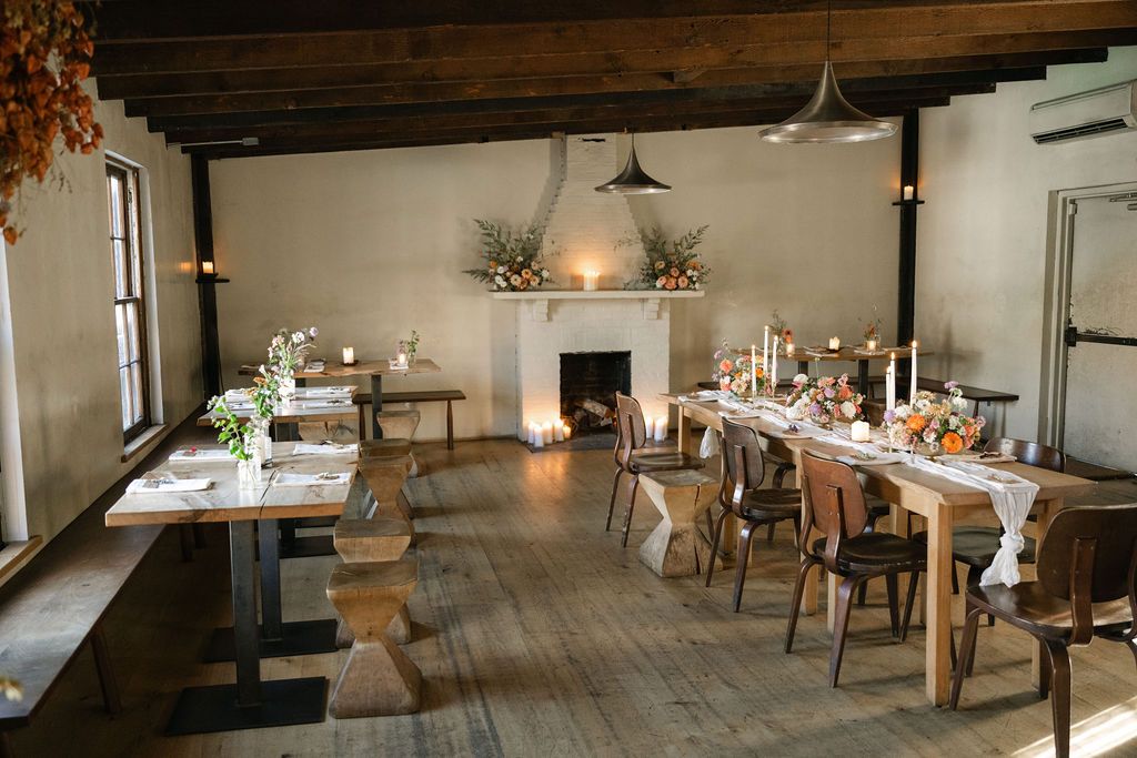 A rustic dining room with wooden tables, chairs and a white fireplace decorated for an event.