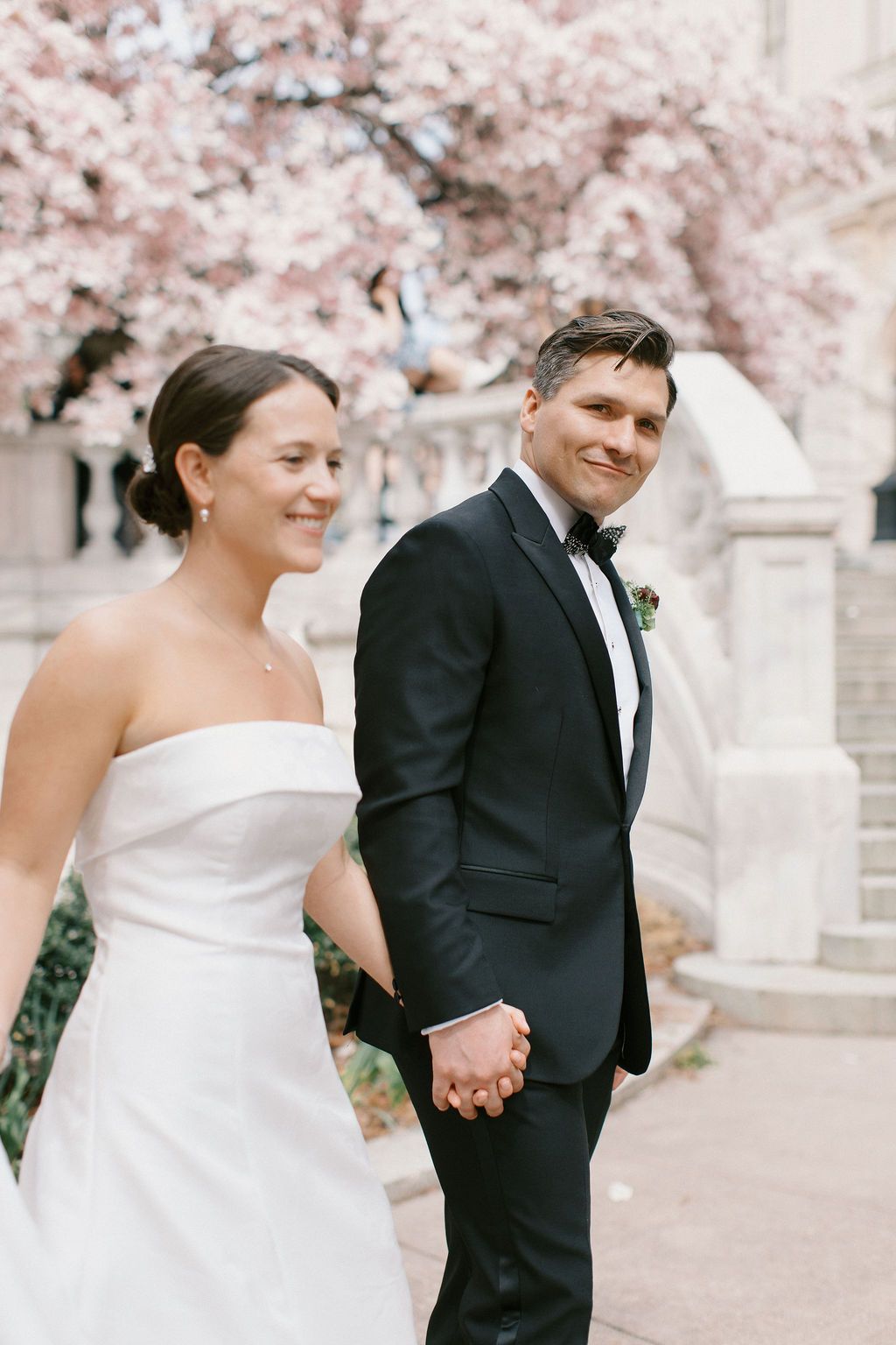 Couple walks hand in hand under cherry blossoms in wedding attire.