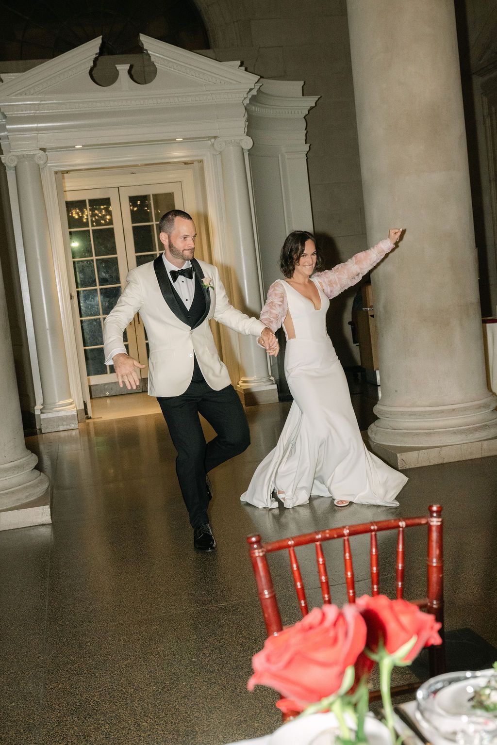 A couple dances joyfully in an elegant hallway with white columns and red chairs visible in the foreground.