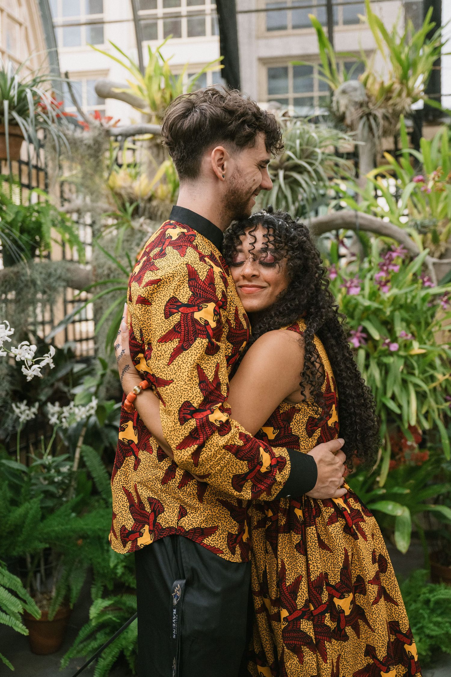 Couple in matching African print outfits embracing in a lush indoor garden with tropical plants.