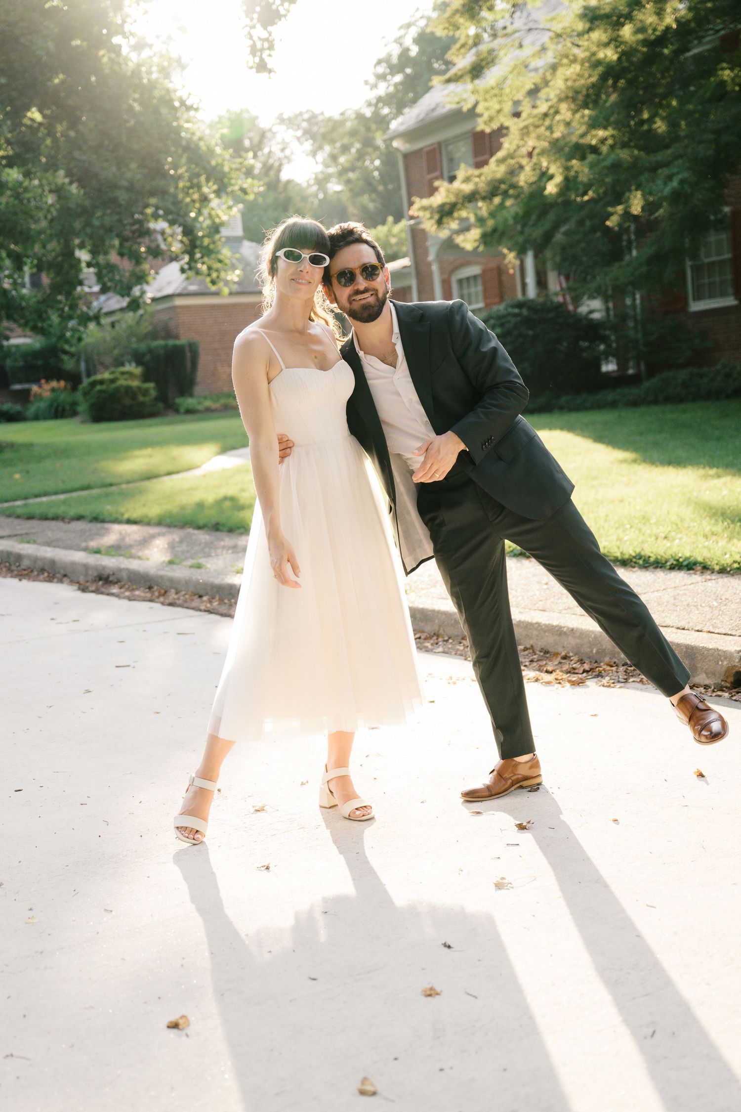 A couple in wedding attire poses playfully on a sunlit sidewalk with trees and houses in the background.