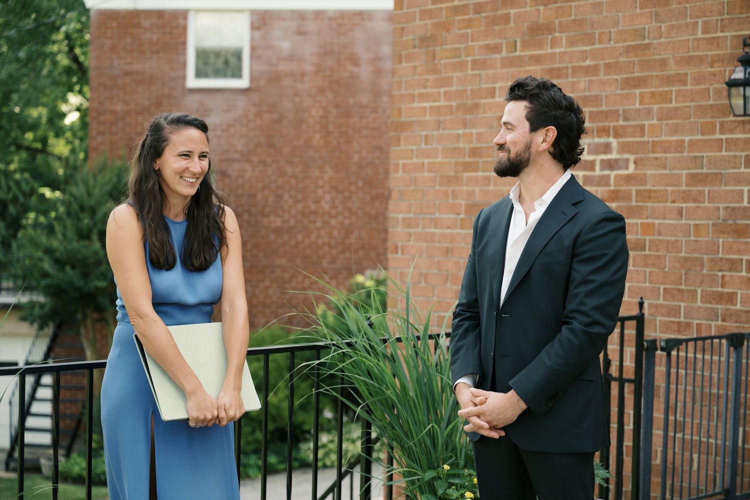 Couple in formal attire standing by brick building exterior during wedding photoshoot.