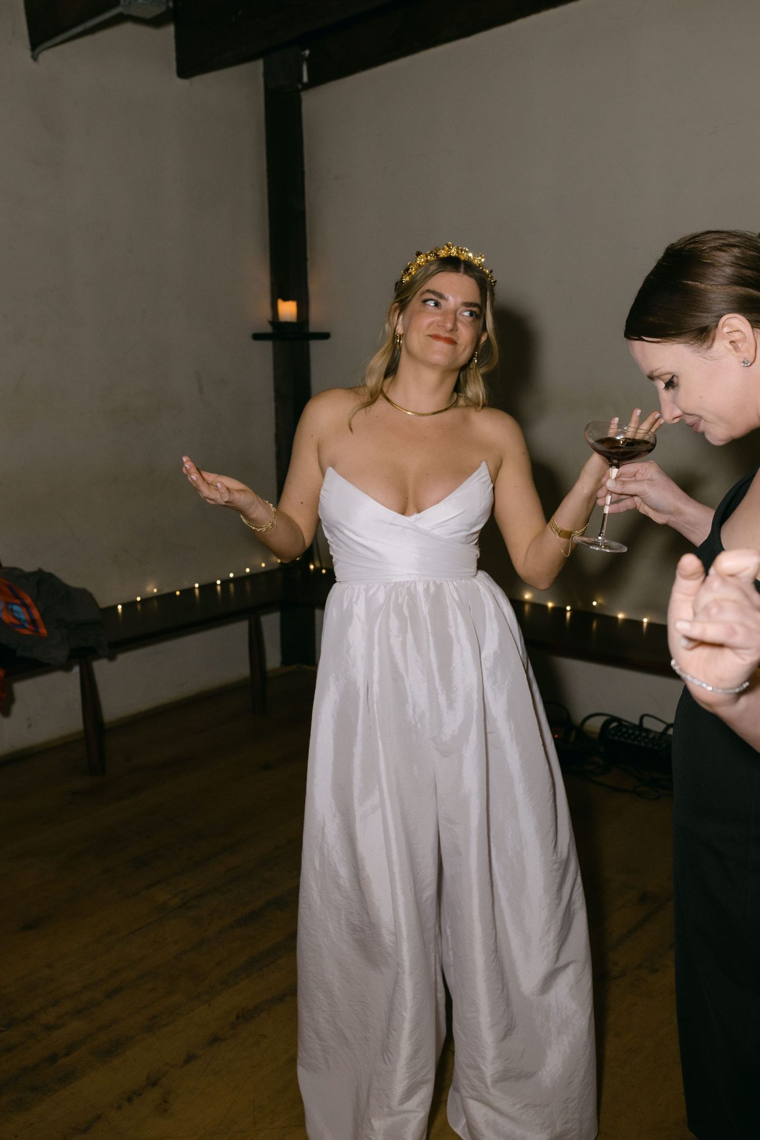 Someone wearing a white strapless wedding jumpsuit with a gold crown making a gesture at an indoor reception.