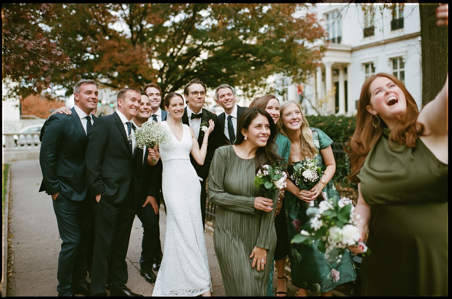 Wedding party gathered outdoors on an autumn day with trees in background.