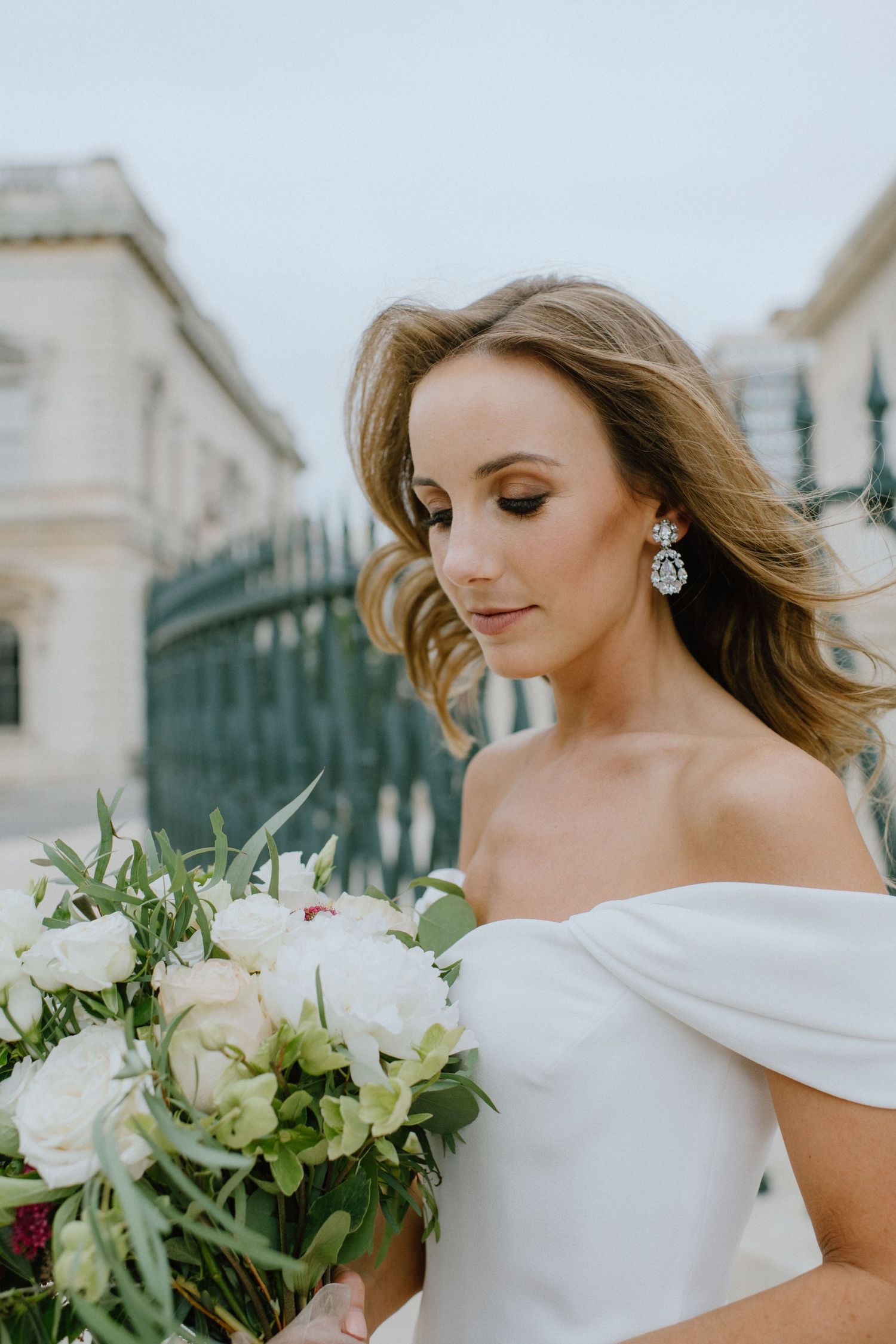 An elegant bridal portrait features a white off-shoulder gown and sparkling earrings with a lush white bouquet.
