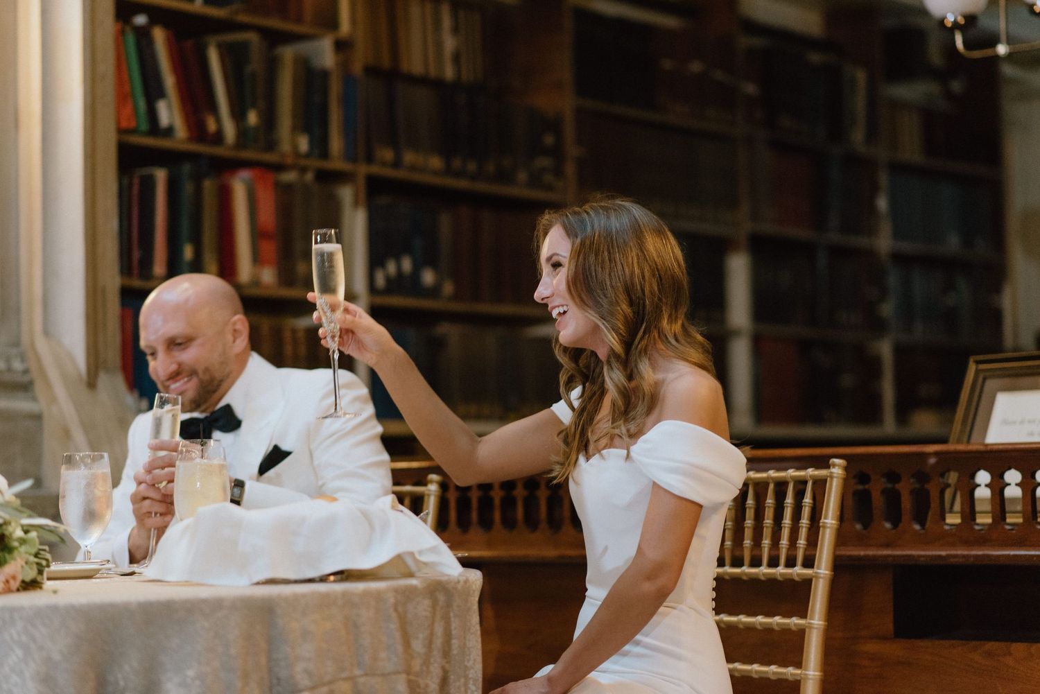 Bride in white dress raises champagne glass for a toast in a historic library setting.