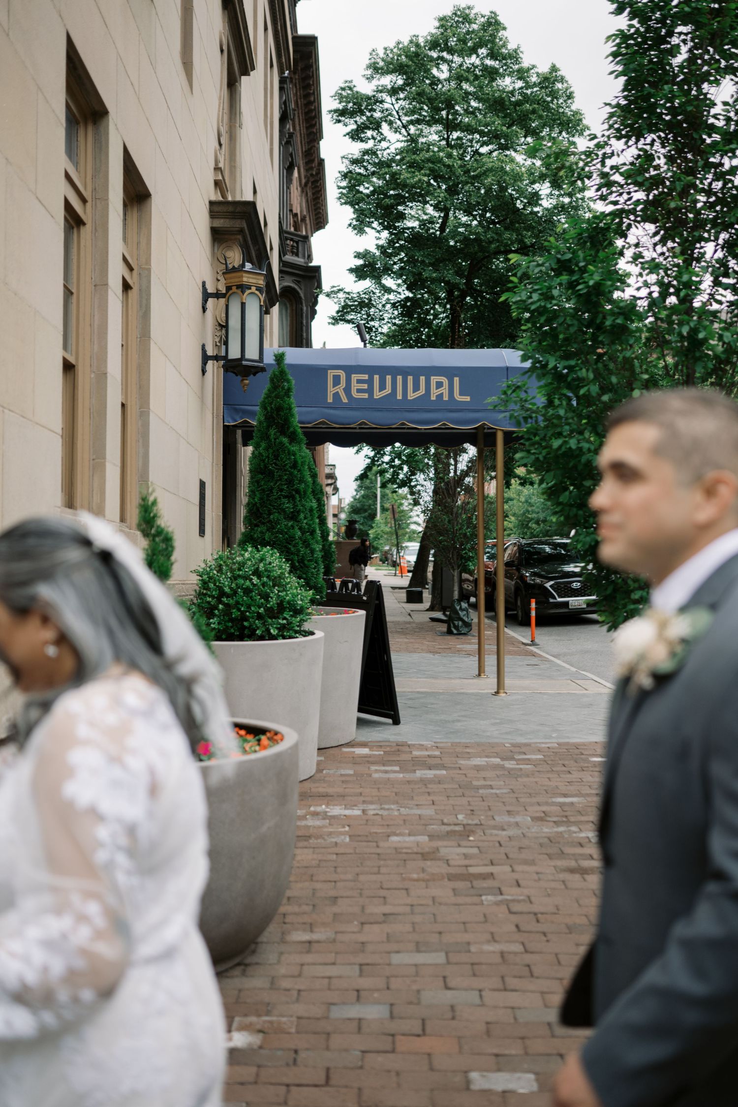 A wedding couple stands on a brick sidewalk near an urban storefront with a 'Revival' sign overhead.