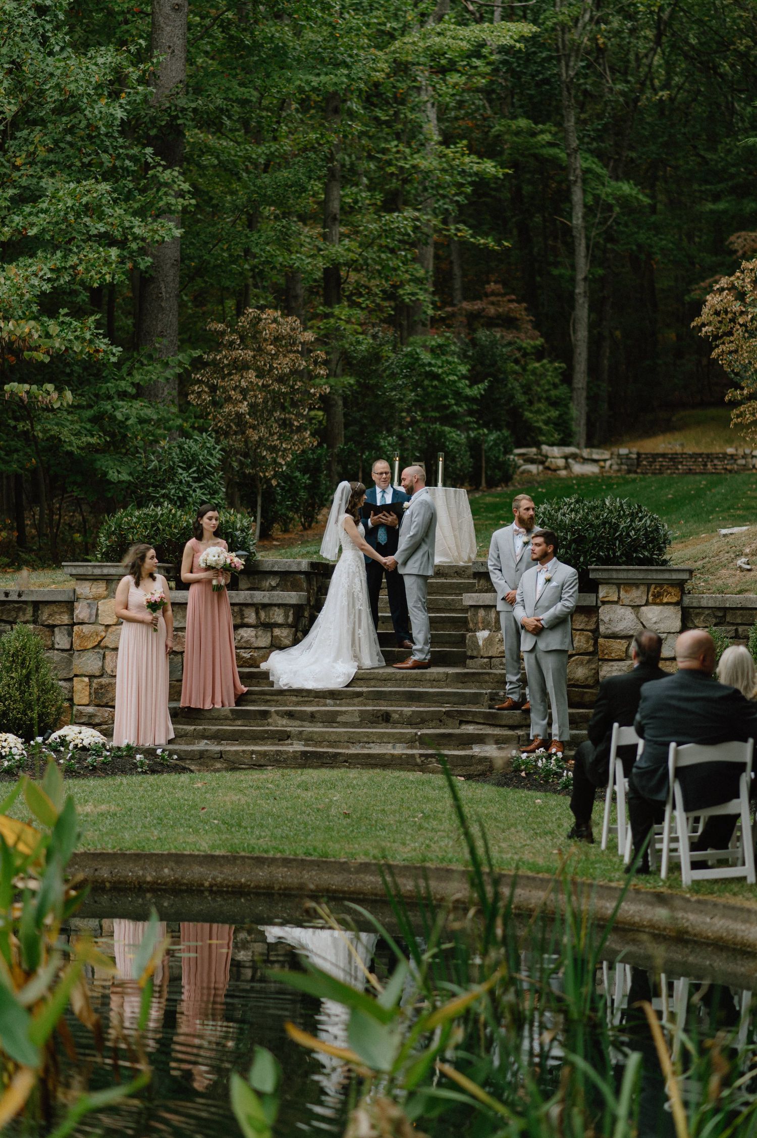 Outdoor wedding ceremony by a pond with bridal party in pink dresses on stone steps.