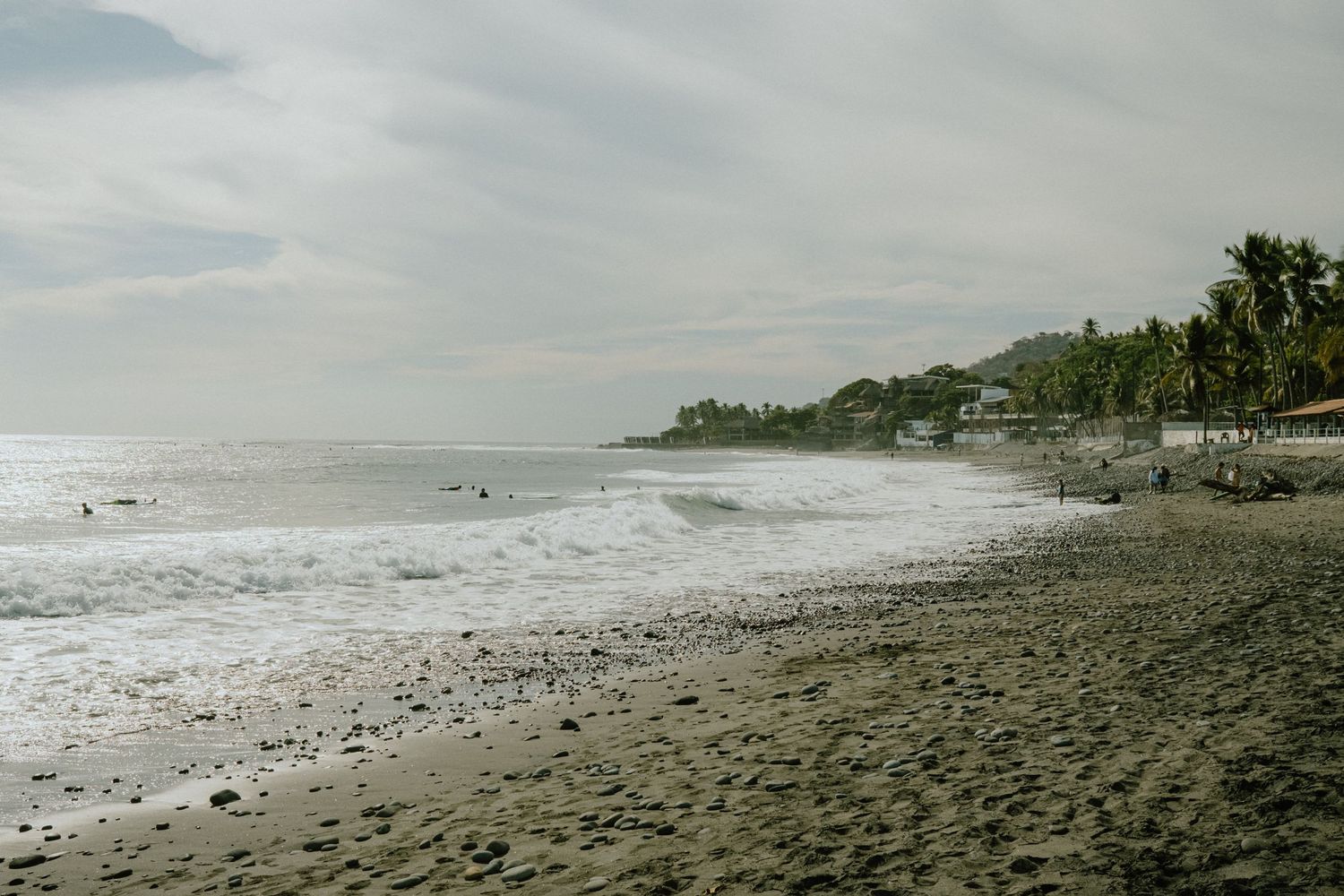 A scenic beach with dark sand and waves rolling in under cloudy skies with palm trees in the distance.