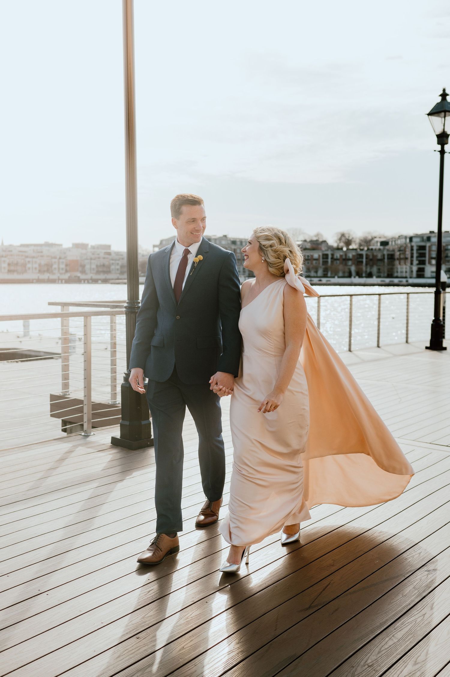 Couple walks hand in hand on a waterfront boardwalk during golden hour, with formal attire and flowing dress cape.