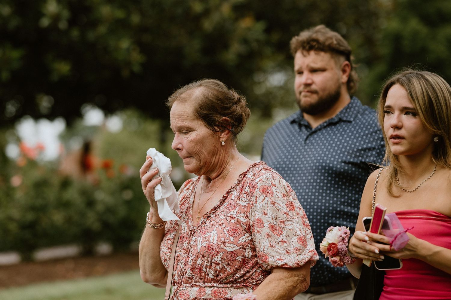 Wedding guests share emotional reactions while drinking from glasses in an outdoor garden setting.