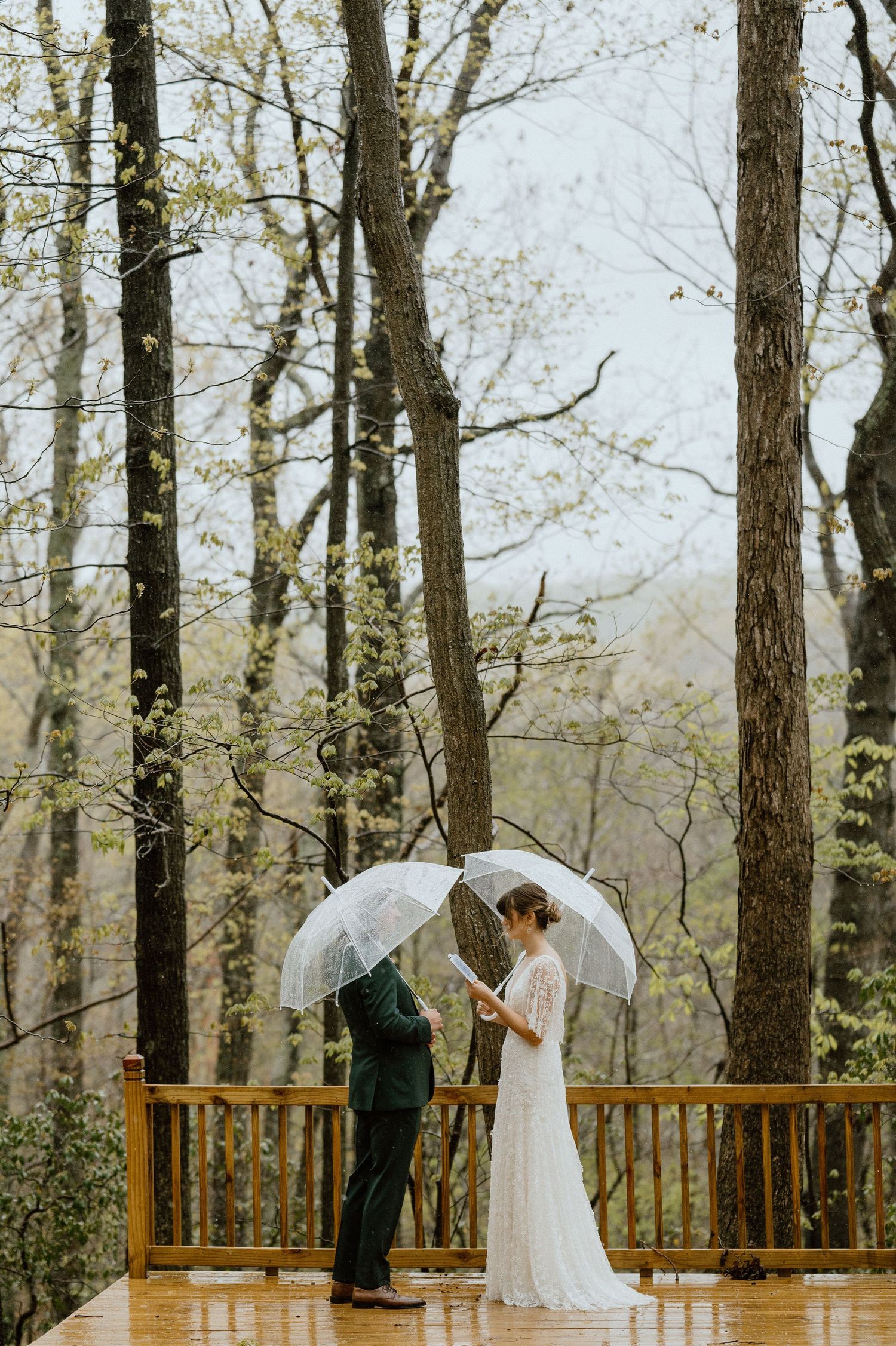 Wedding couple sharing an intimate moment under a clear umbrella on a rainy deck.