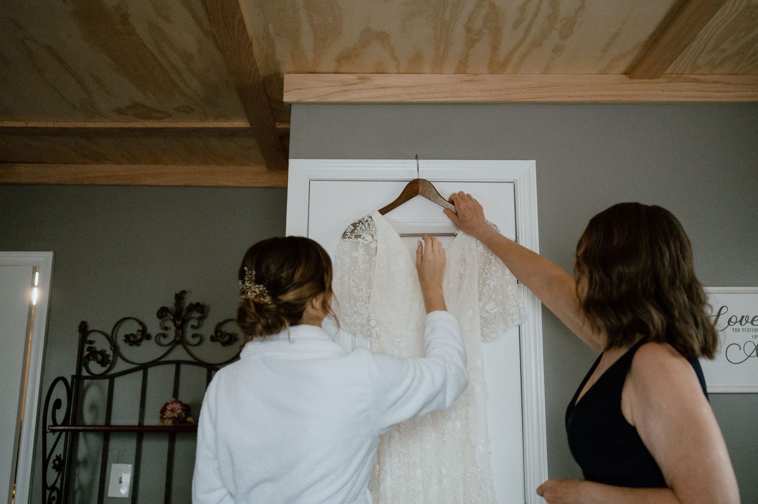 Two people reaching up to handle a white wedding dress hanging on a door.