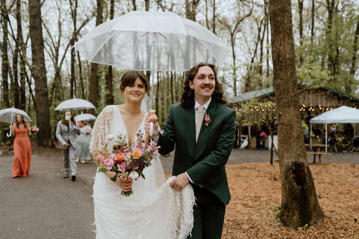 A couple walks under clear umbrellas during their rainy woodland wedding day.