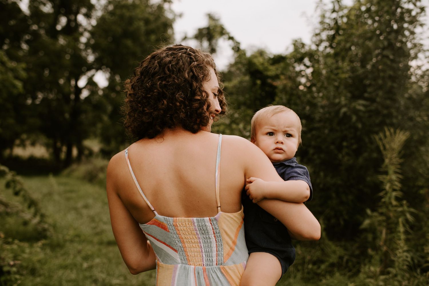 Woman in striped dress holding baby outdoors in green summer garden setting.