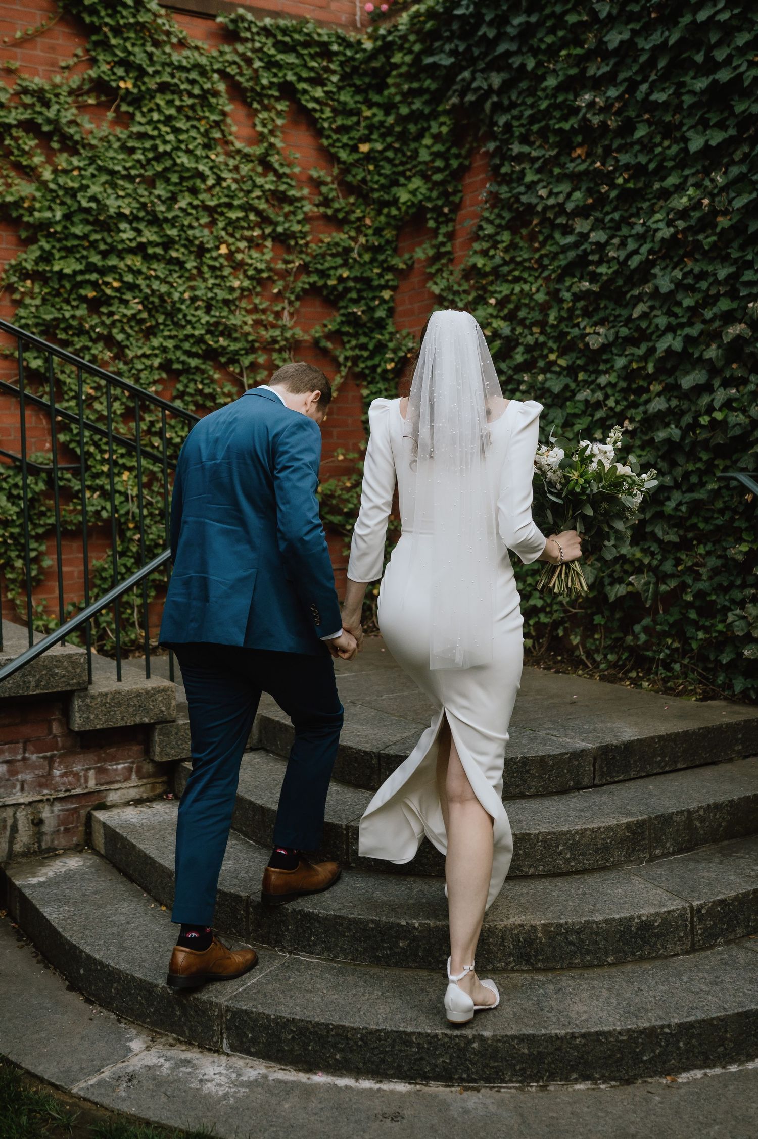 A couple in wedding attire ascends brick steps with ivy-covered walls in the background.