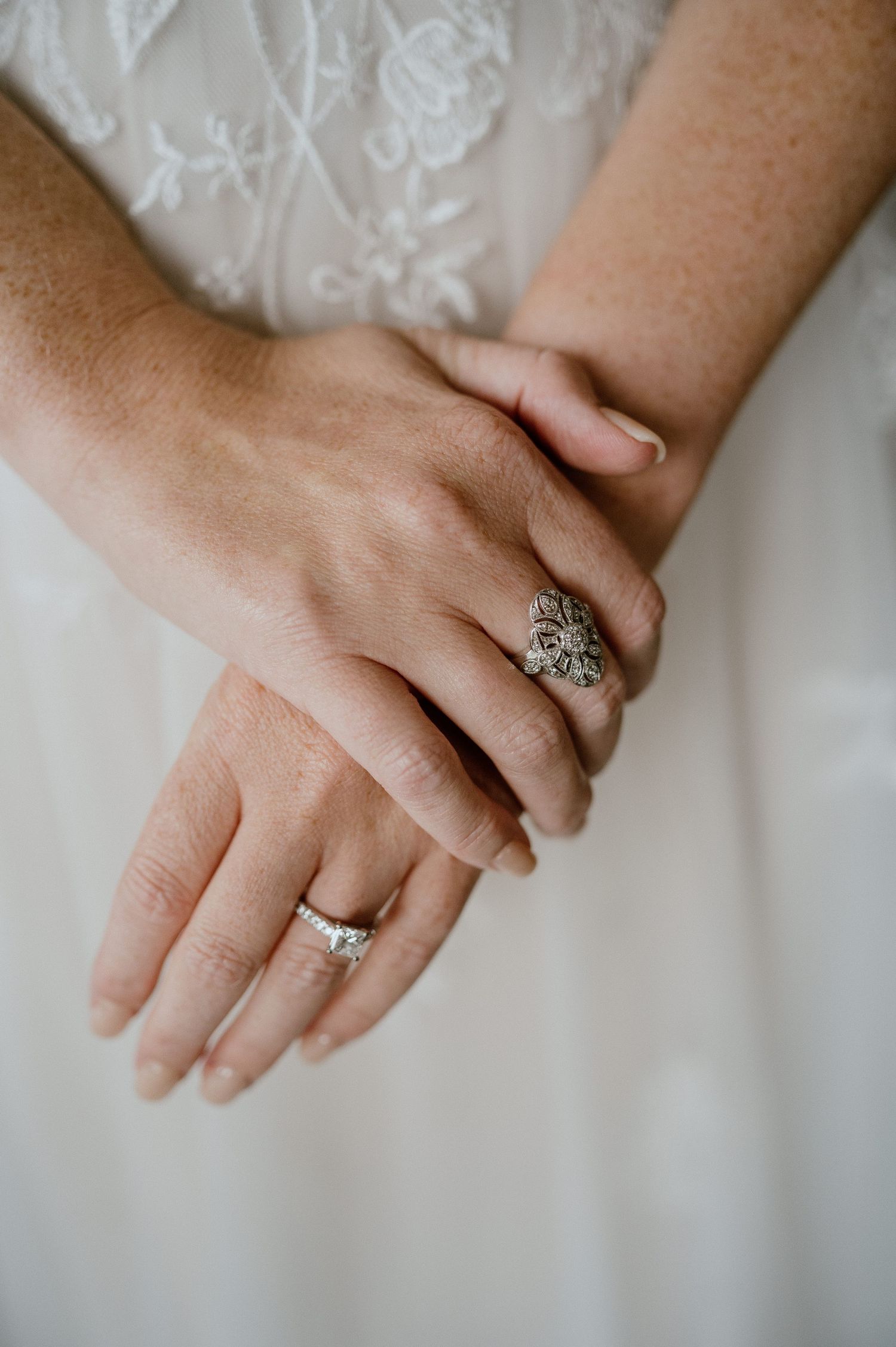 Close-up of hands wearing elegant vintage and modern wedding rings against a white lace dress backdrop.