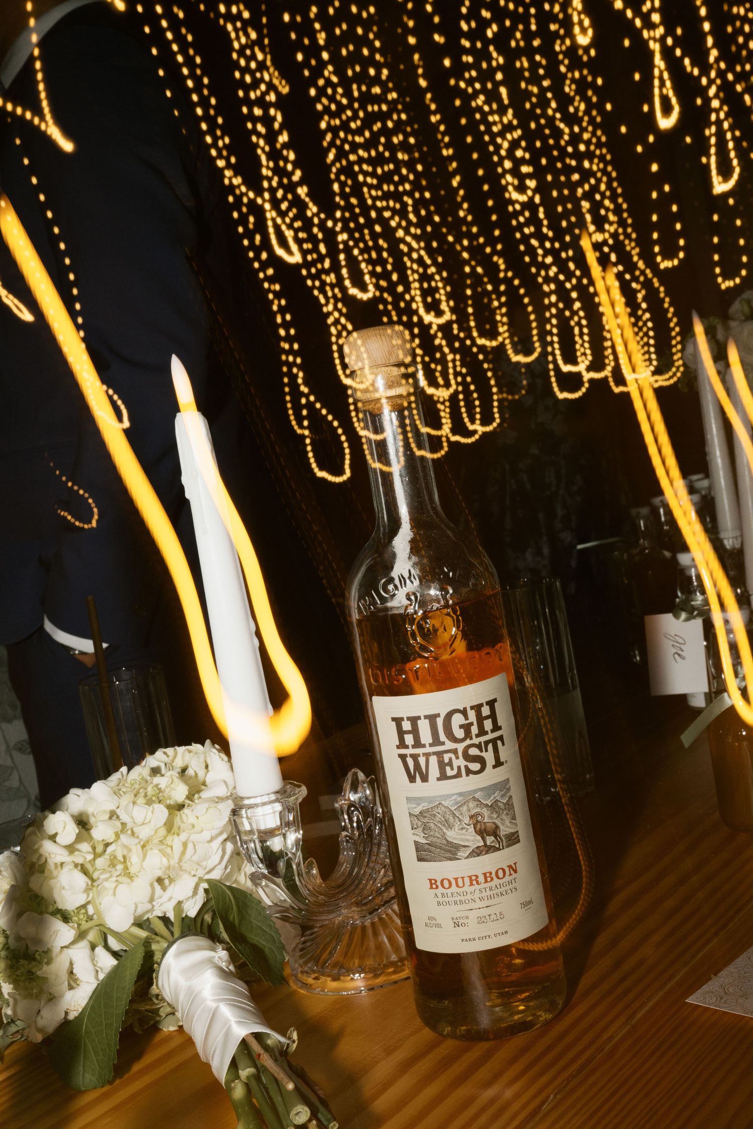 Wedding reception decor with illuminated string lights and bourbon bottles on wooden table.