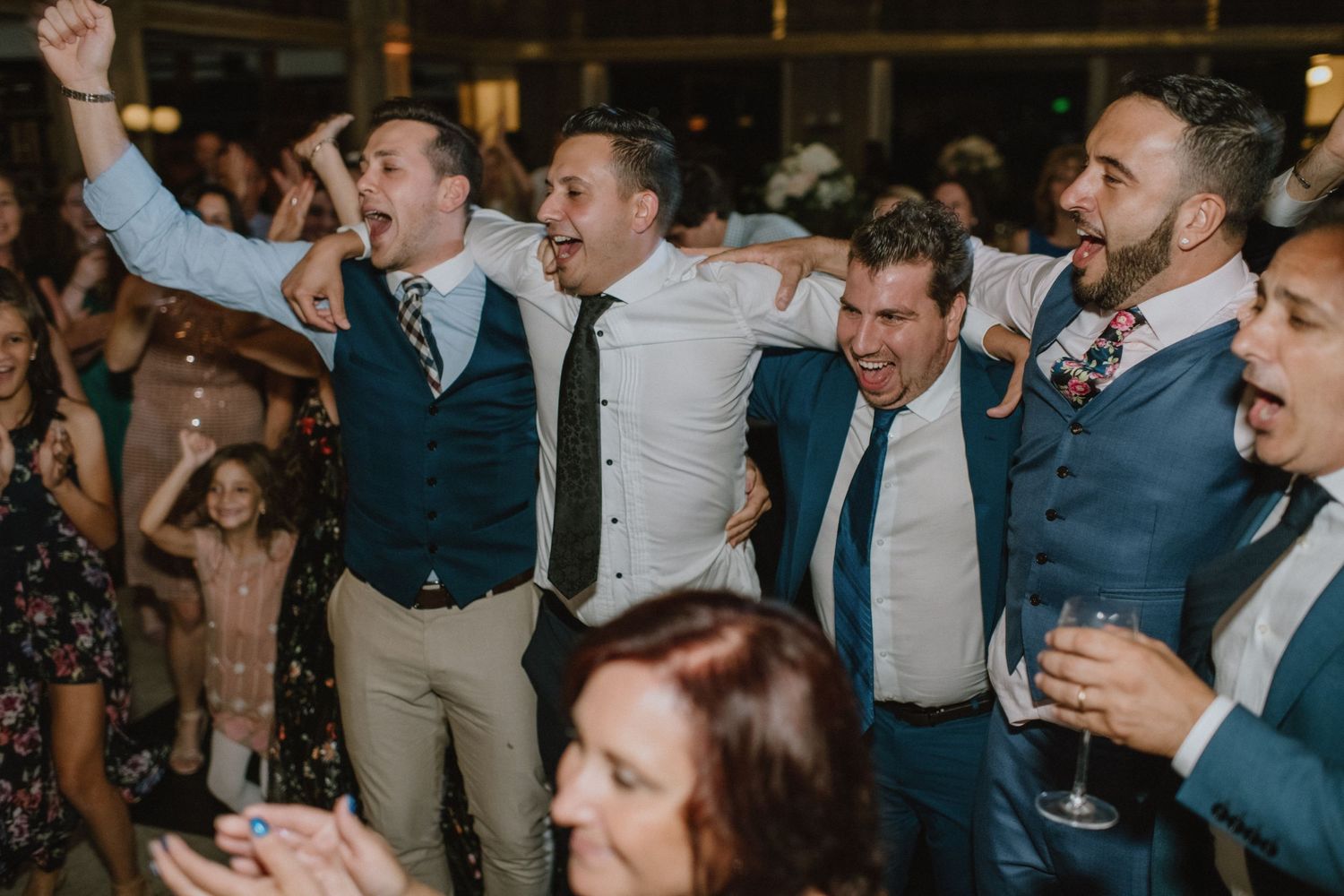 Group of guests in formal attire celebrating enthusiastically at an indoor wedding reception.
