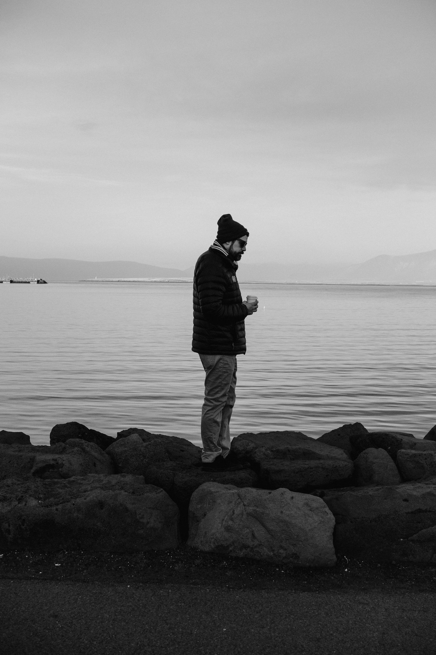 A silhouetted figure stands on rocky shoreline overlooking a calm body of water in black and white.