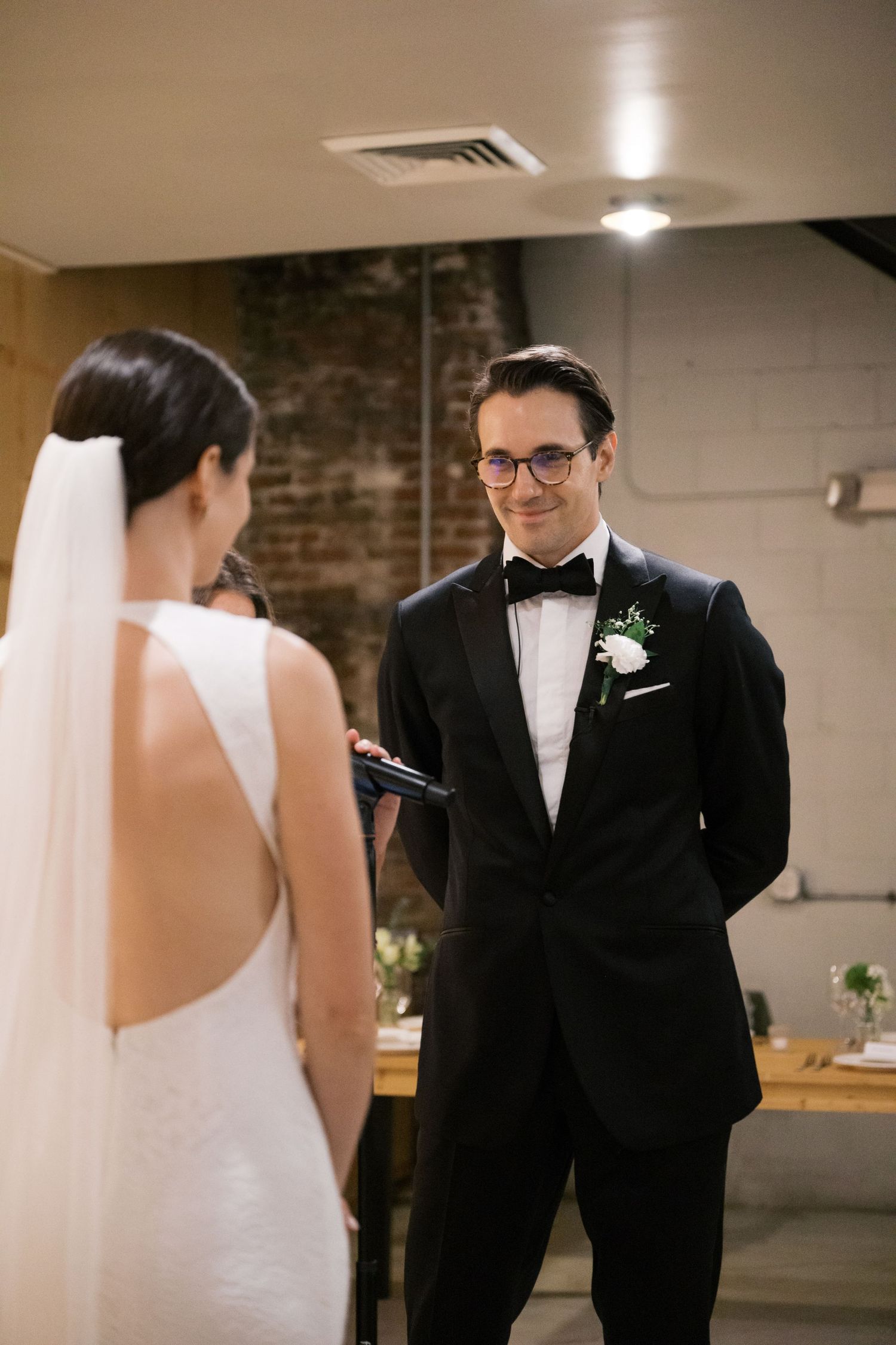 Bride in keyhole back dress and groom in tuxedo exchanging vows during indoor wedding ceremony.