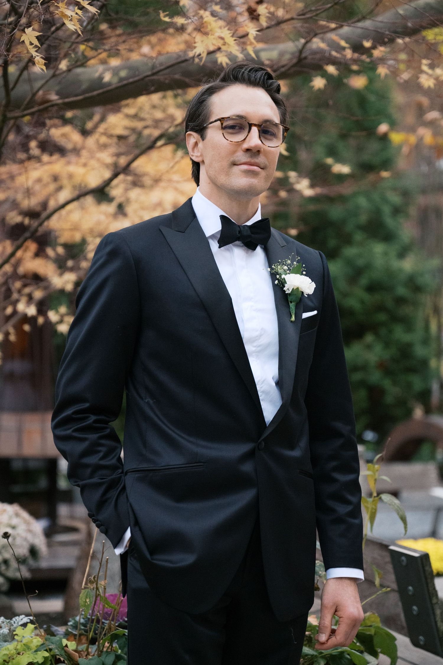 Groom in classic tuxedo and bow tie poses against autumn foliage.