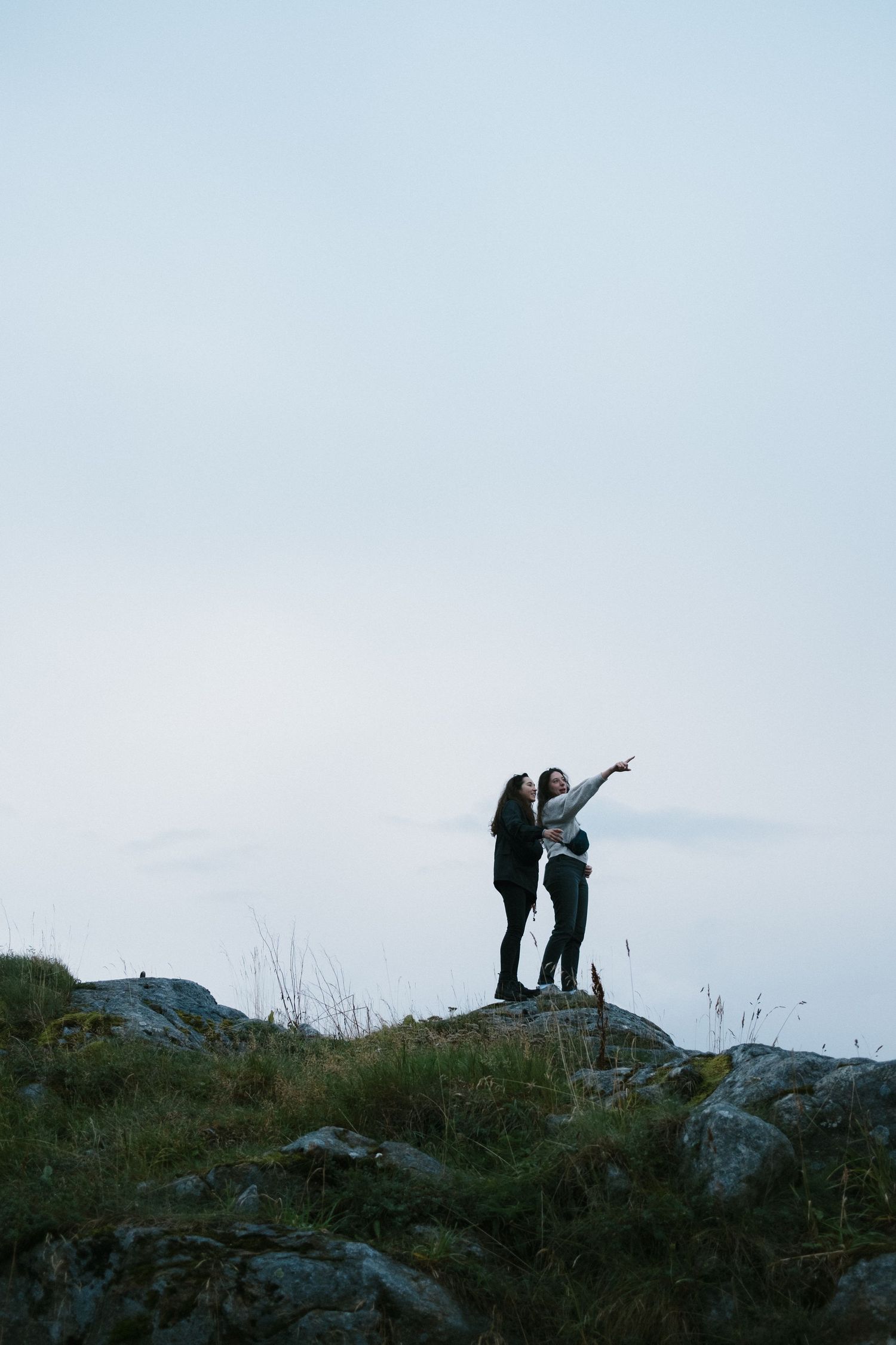 Two silhouettes stand together on a hilltop against cloudy sky at dusk.