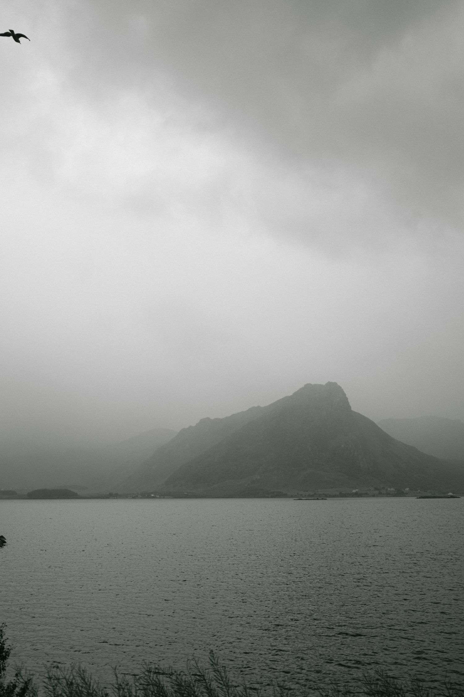 Misty mountain peak rises dramatically over a calm lake on an overcast day.